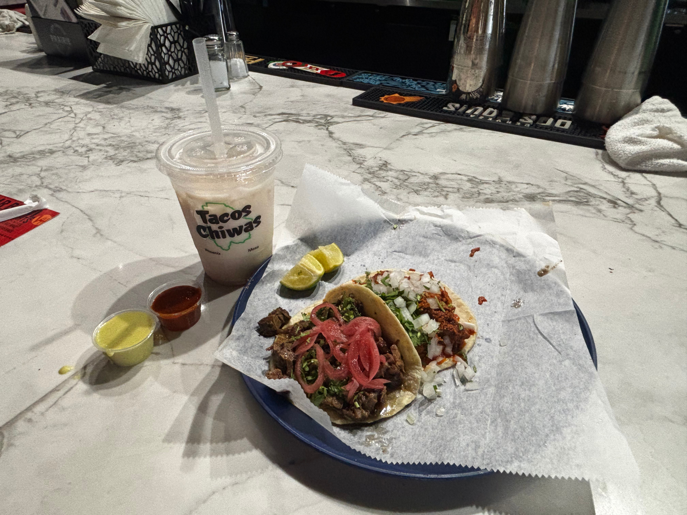 Marble bar counter with a blue plate of two tacos topped with cilantro and onions, lime wedges, and sauces; an iced drink in a "Tacos Chiwas" cup sits nearby, bar taps in the background.