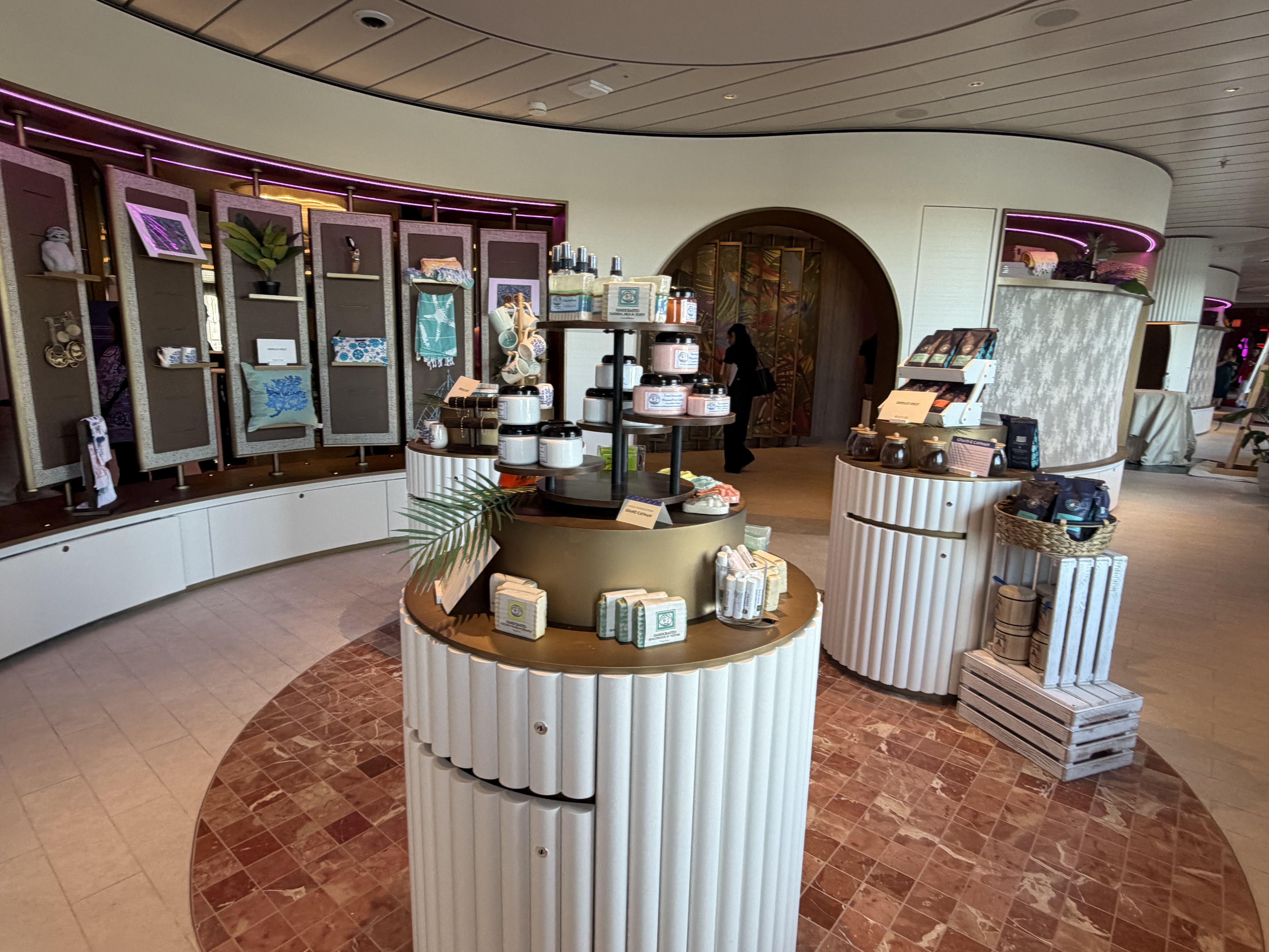 Interior of a boutique with round white display tables holding skincare and wellness products, decorative wall panels with textiles and art, and a curved ceiling with purple lighting.