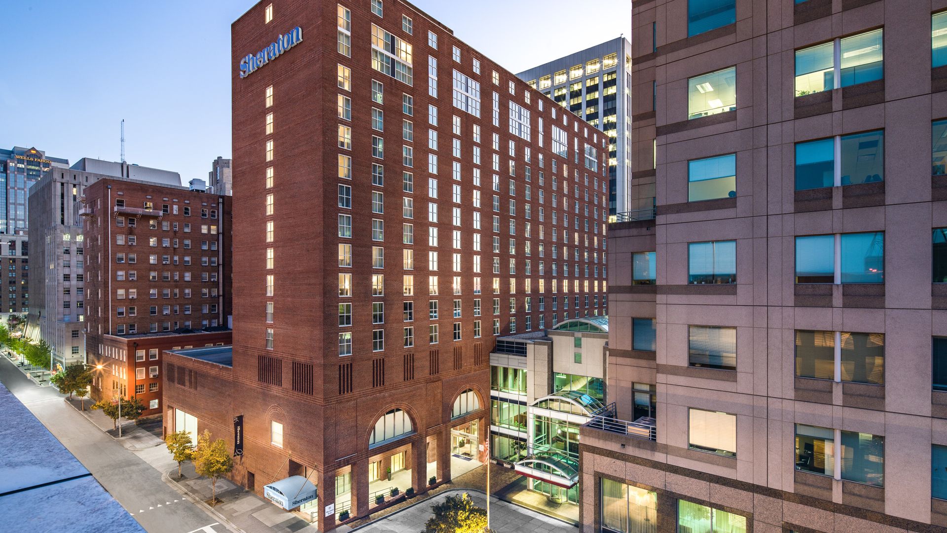Aerial view of a city block with a tall red-brick Sheraton hotel and its sign, white-framed windows, adjacent modern glass towers, a glass canopy atrium, and a tree-lined street.