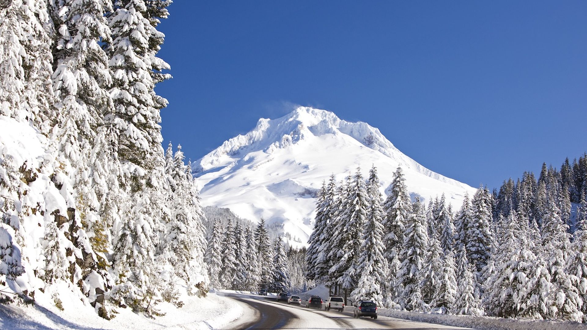 A photo of a white-capped mountain peak with a snow-covered street with cars driving up toward it.