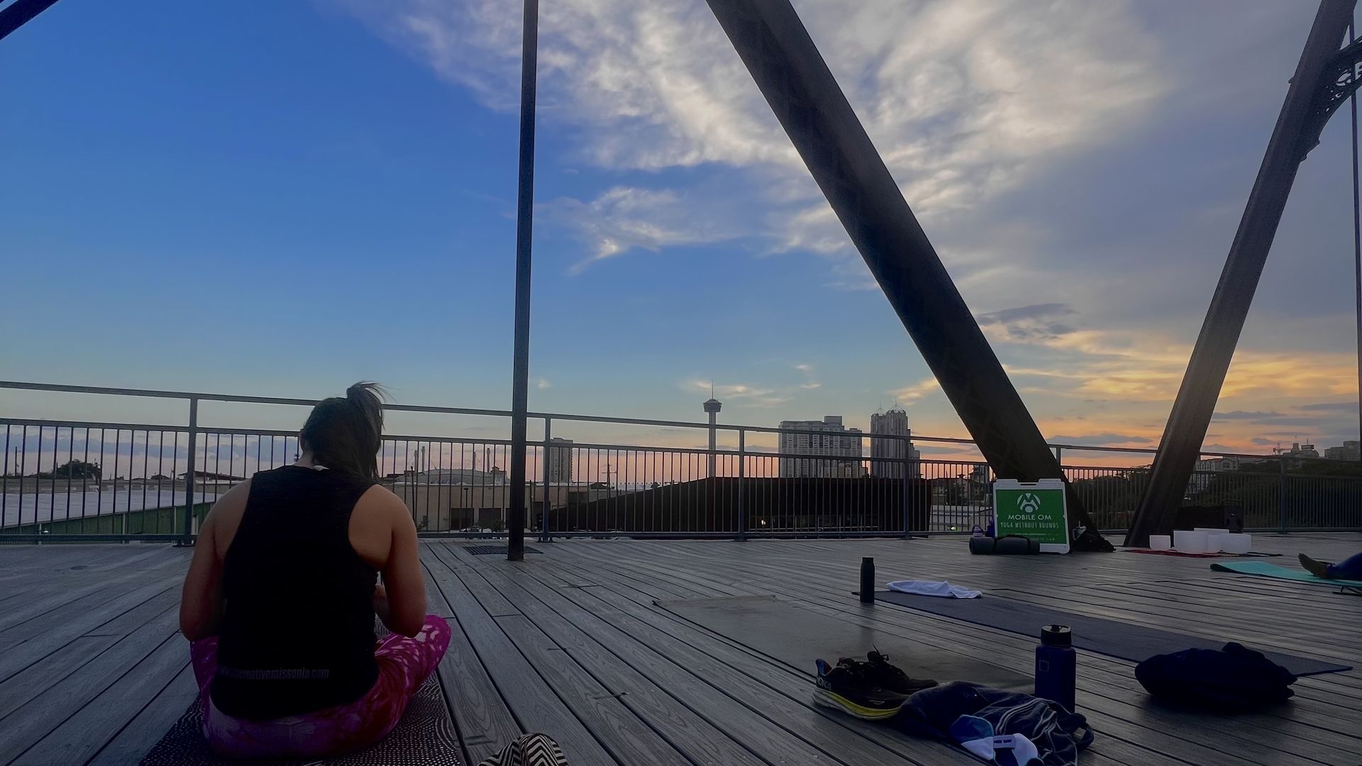 Person sitting on a yoga mat on a rooftop deck at sunset, with city buildings and a tower in the background. Yoga mats, shoes, and water bottles are scattered around.