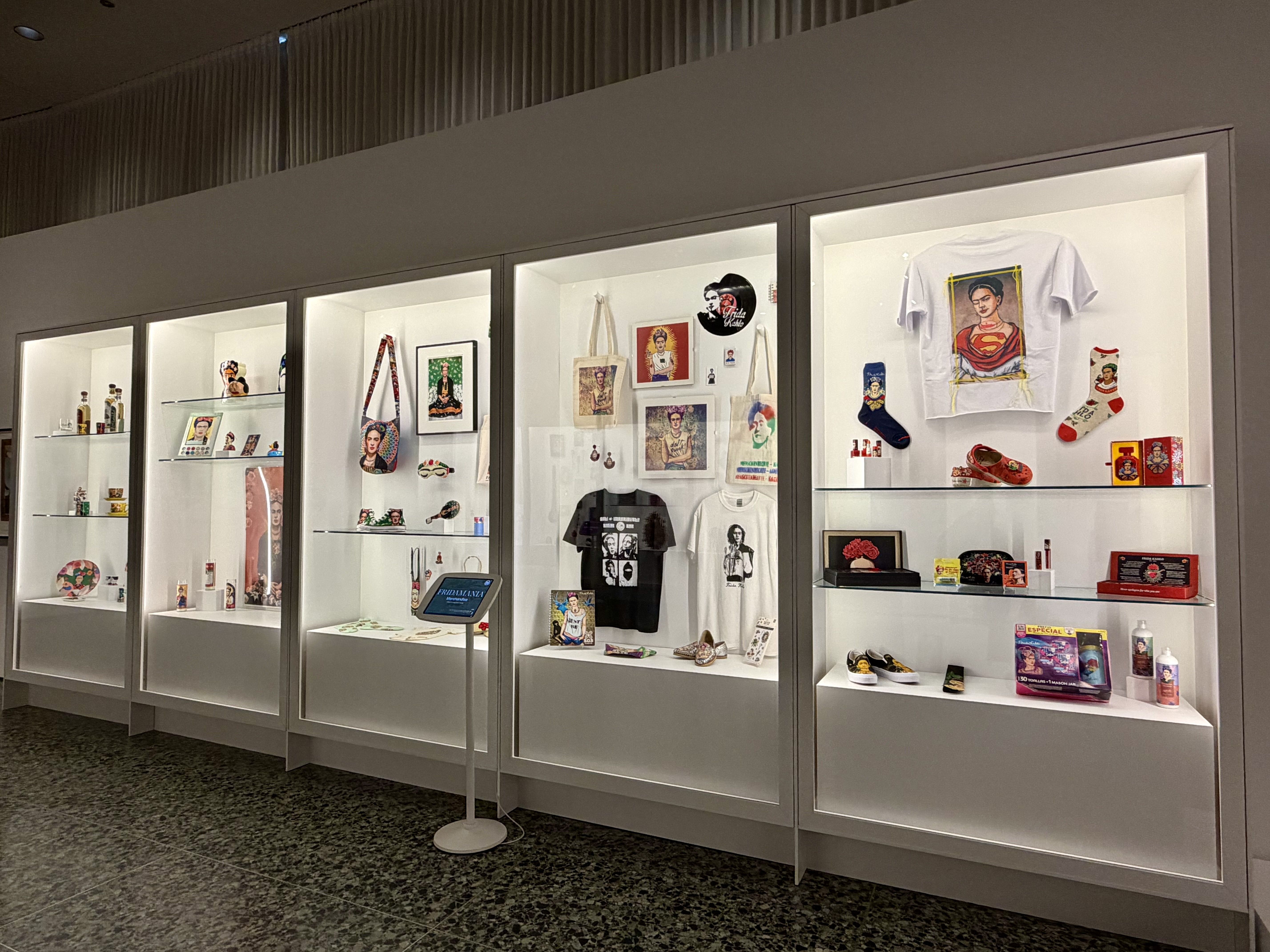 A museum display case showing various items featuring Frida Kahlo, including T-shirts, bags, shoes, socks, portraits, and decorative objects on white lit shelves.