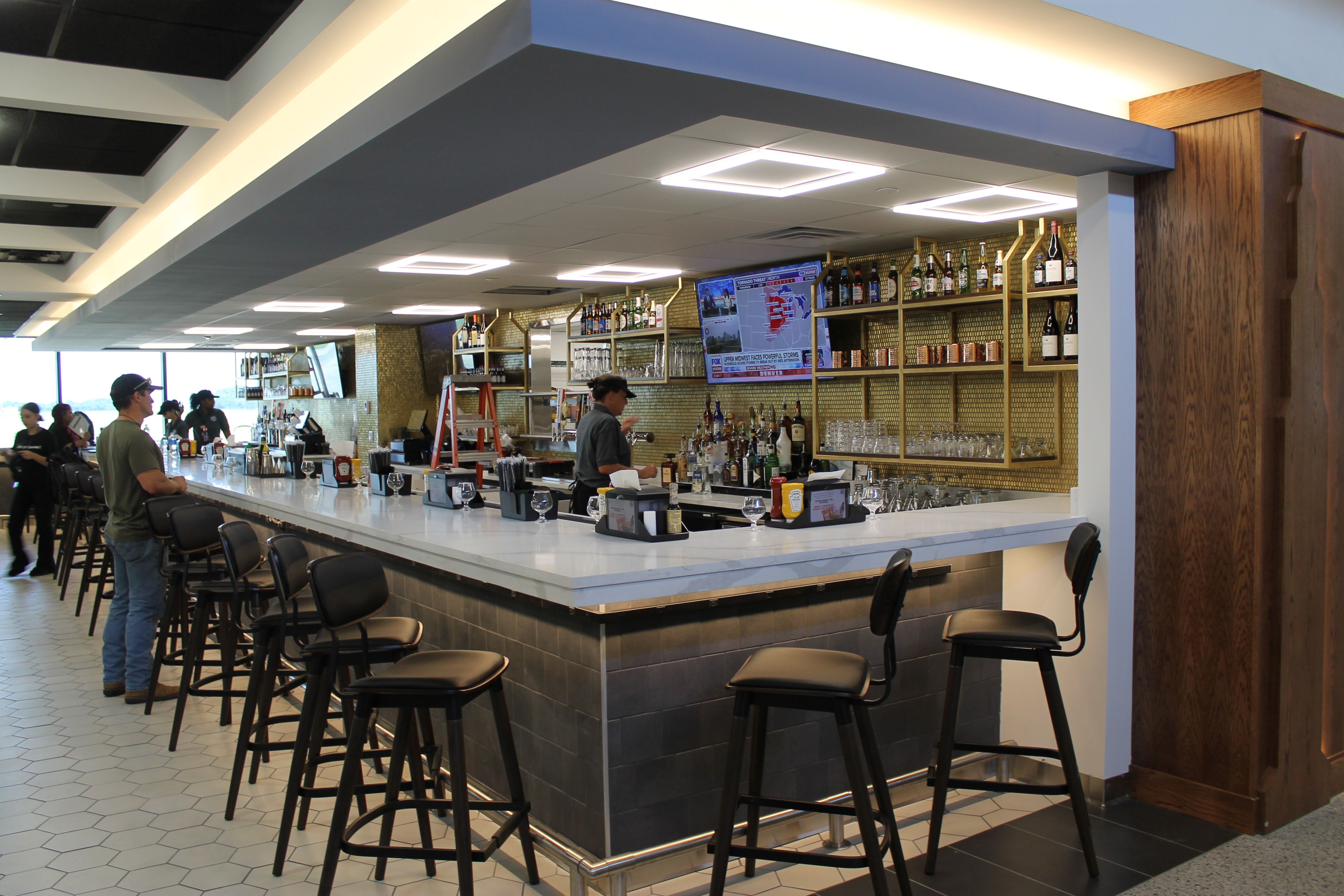 Bright modern bar with a long white counter and black stools. Gold shelving holds bottles; a TV is mounted above the shelves. A few patrons stand and chat near the bar.