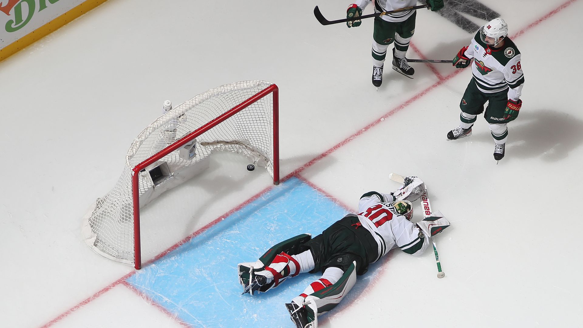 Top-down view of a hockey goal Jesper Wallstedt: he's on the blue crease near the net, two players in green-and-white jerseys nearby, and a puck near the goalie beside the red goal frame.