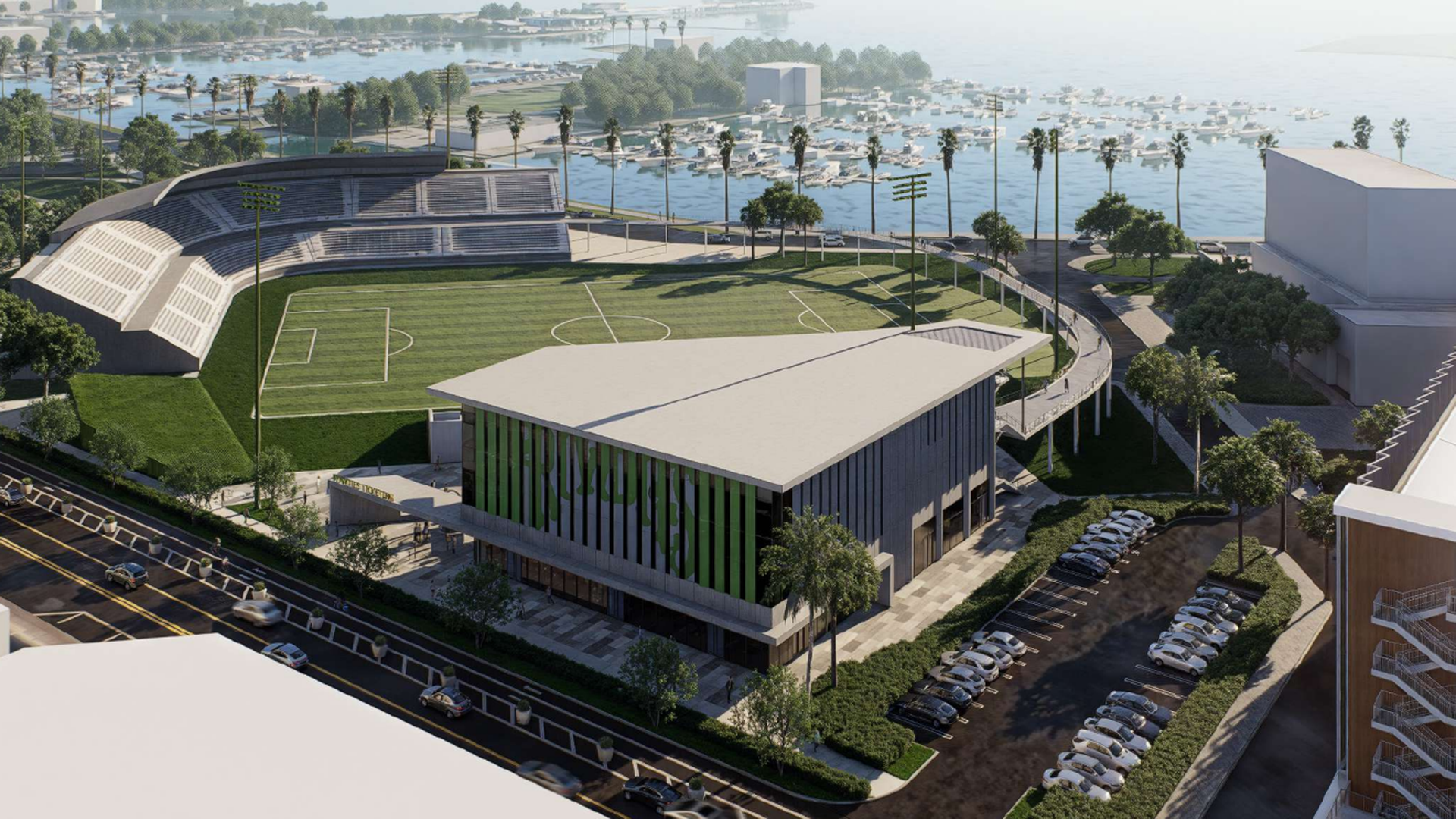 Aerial view of a modern sports complex with a green soccer field, surrounding bleachers, a large building, parking lot, palm trees, and a marina with boats in the background under a clear sky.