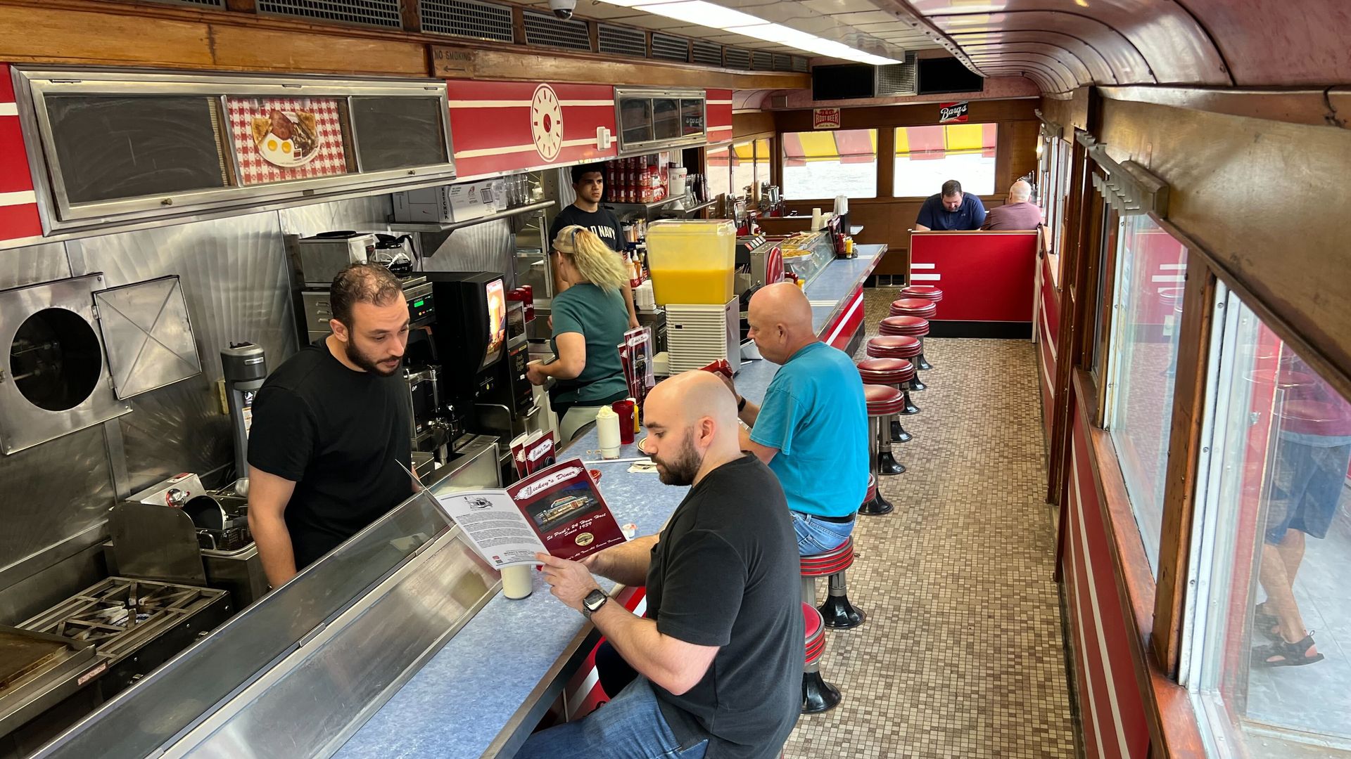 Diners and short-order cooks inside a cramped dining car-style diner with aluminum grills, laminate countertops and wooden trim.