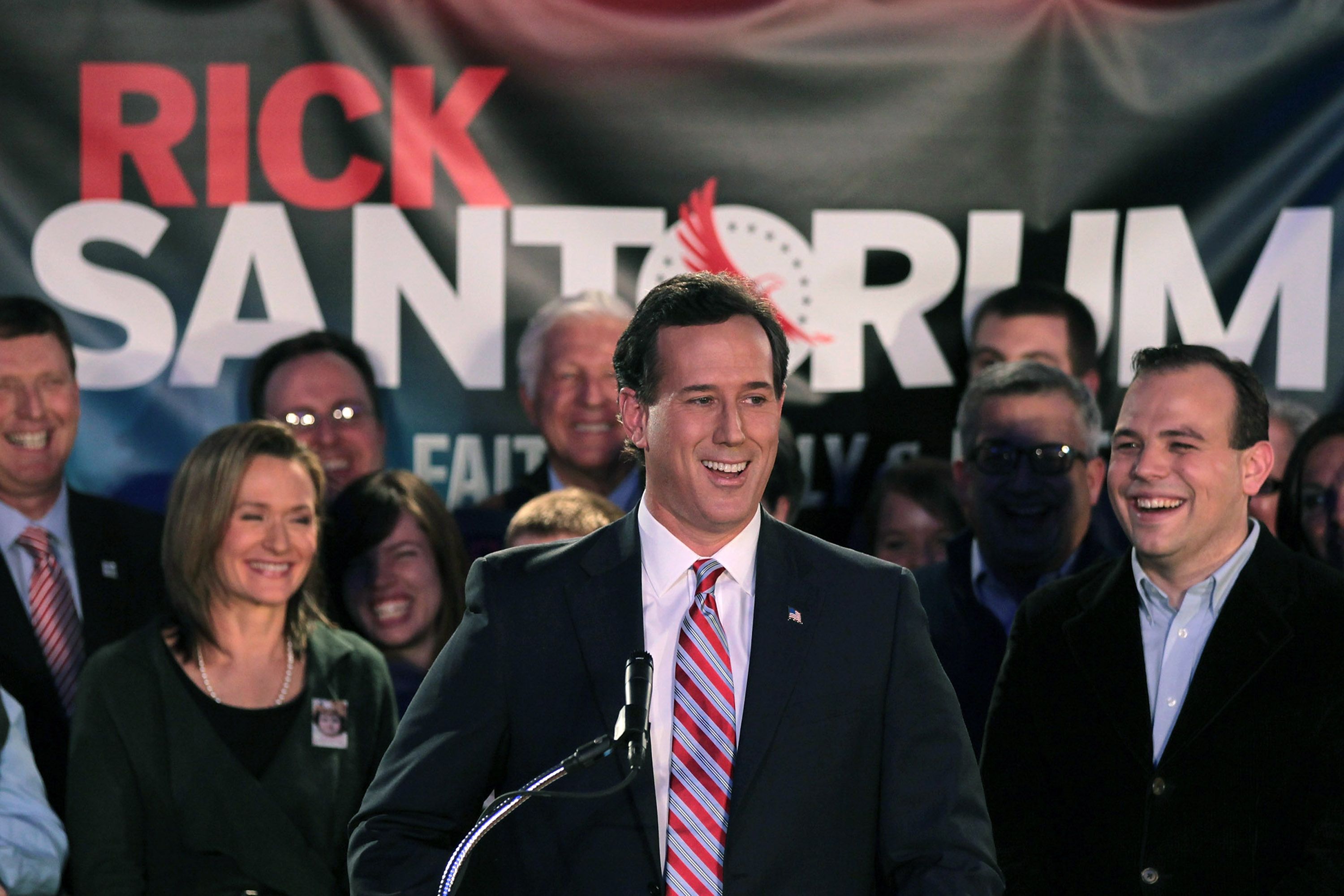 Rick Santorum stands at a microphone with people smiling behind him. A sign says his name in the background.