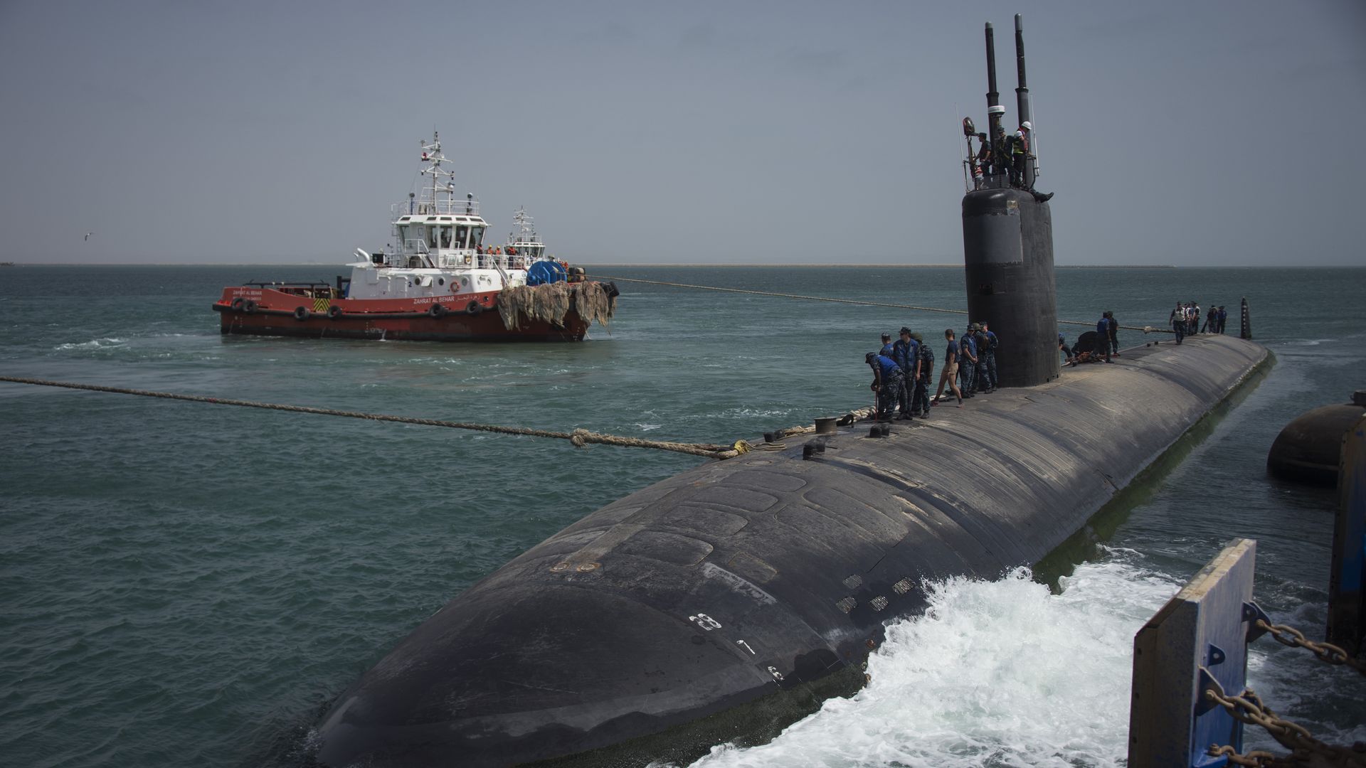 Black submarine being towed by a red-and-white tugboat in a calm blue sea; sailors in blue uniforms line the hull near the conning tower as the tow line links the vessels.