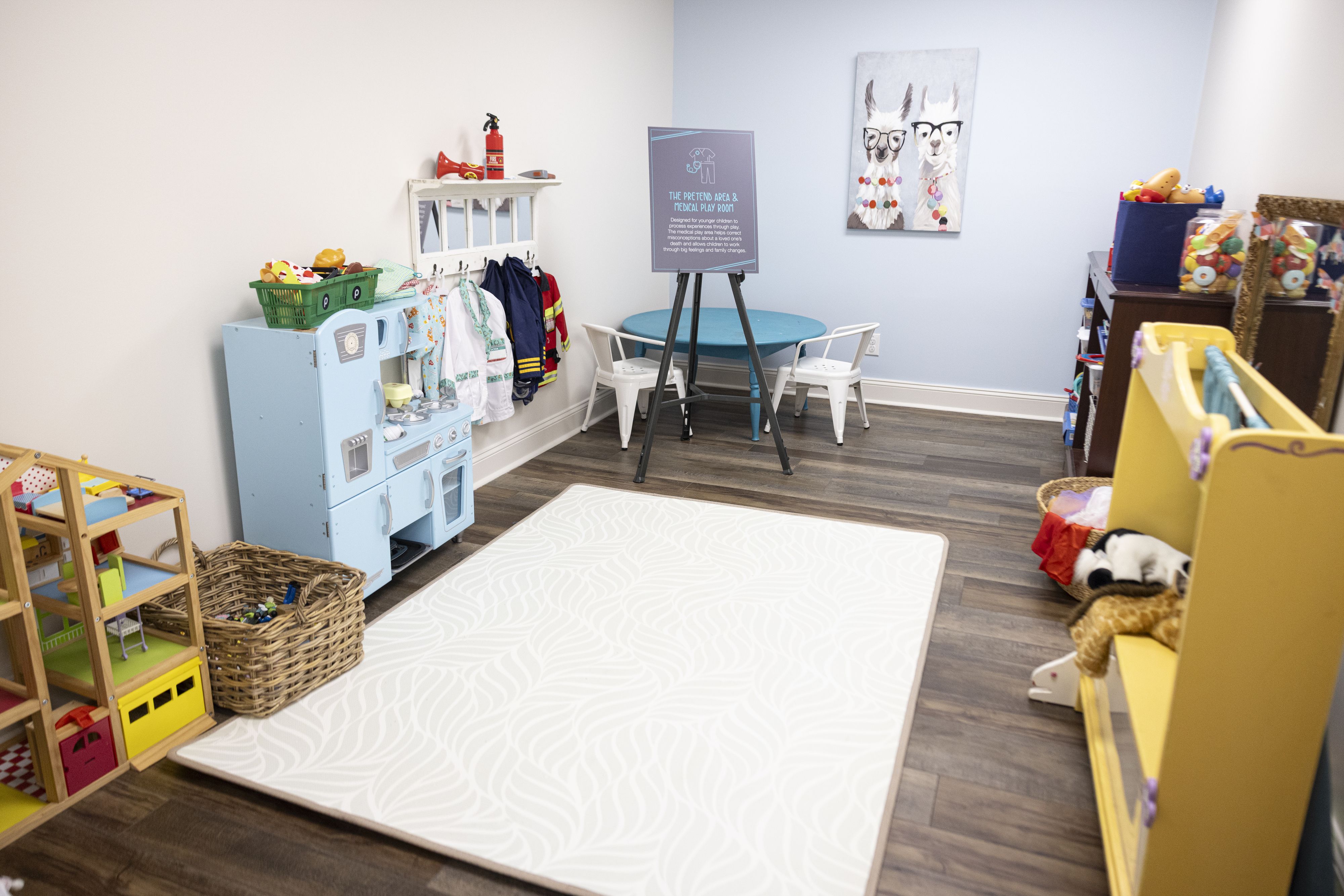 Bright children's playroom with light blue walls, small blue table with white chairs, play kitchen, dress-up clothes, toys, and a rug with a white wavy pattern on dark wooden floor.