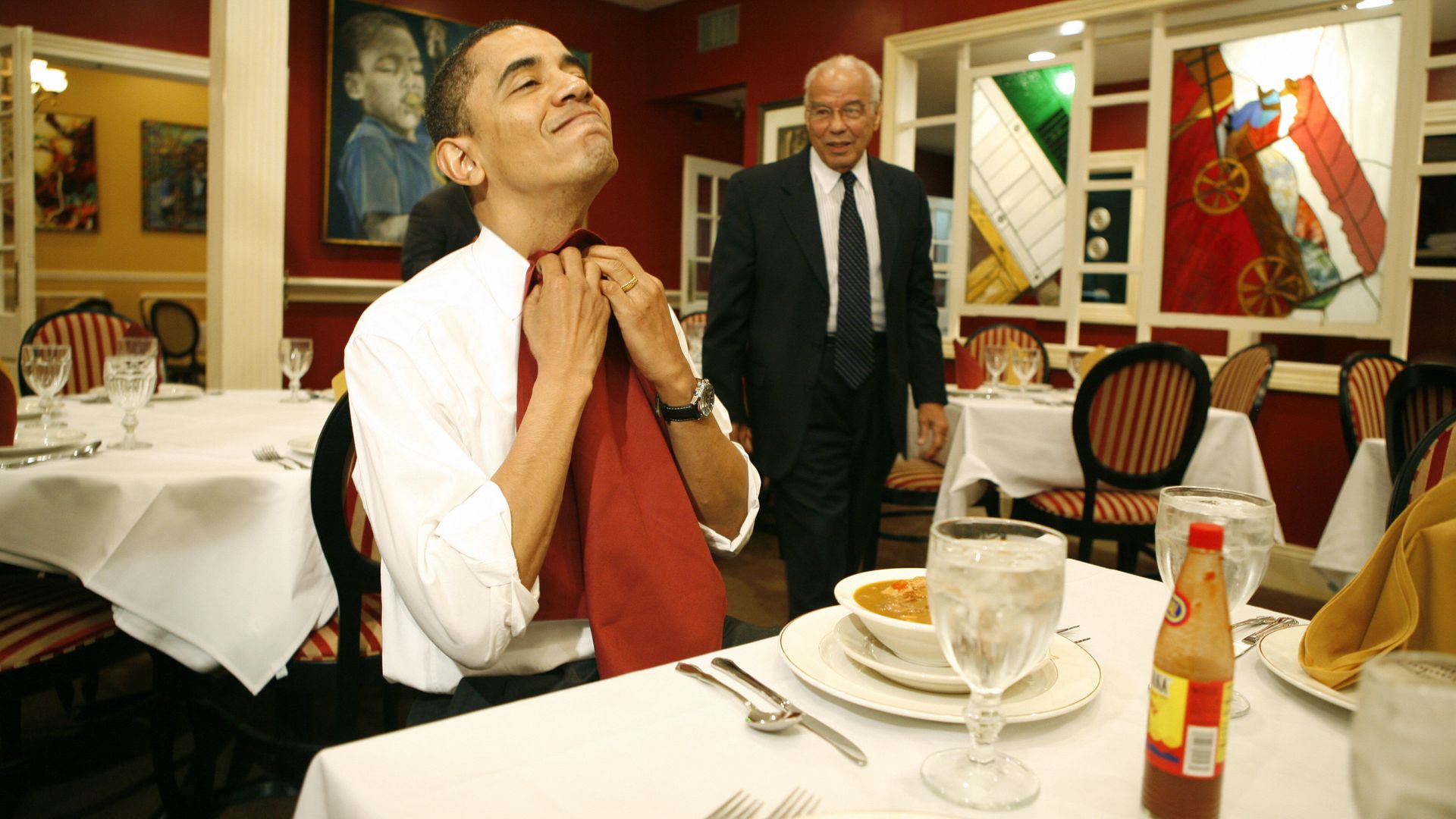 Barack Obama tucks a red cloth napkin into his collar as he sits at a table that's been set with a bowl of gumbo. Dr. Norman Francis looks on as he approaches the table.