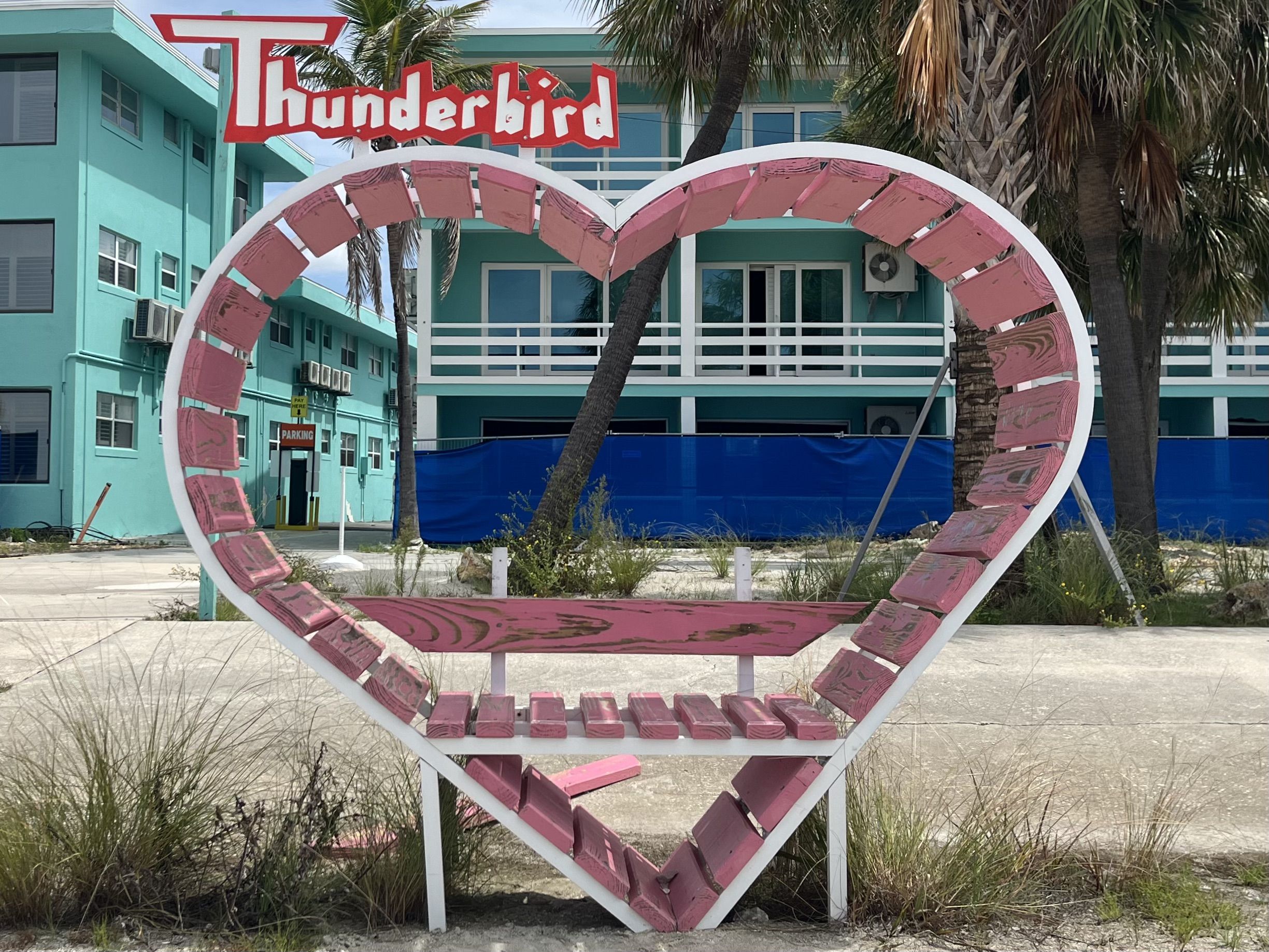 Pink wooden heart-shaped bench with a red and white "Thunderbird" sign above, set outdoors on a concrete surface with turquoise buildings blocked off by a chain-link fence in the background.