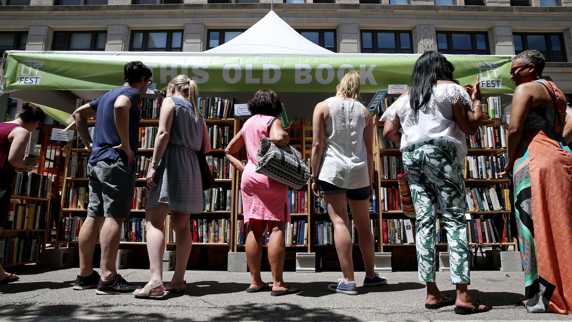 A photo showing seven people outside, looking at books on bookshelves under a tent.