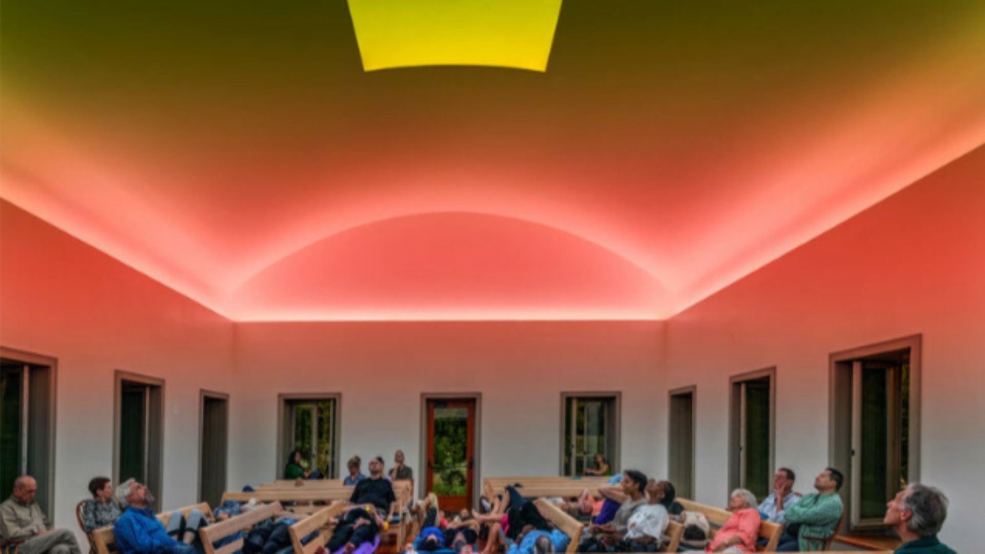 Indoor courtyard bathed in pink-orange ceiling lighting; people sit on wooden benches around a central open area, white walls and tall rectangular windows on the sides.