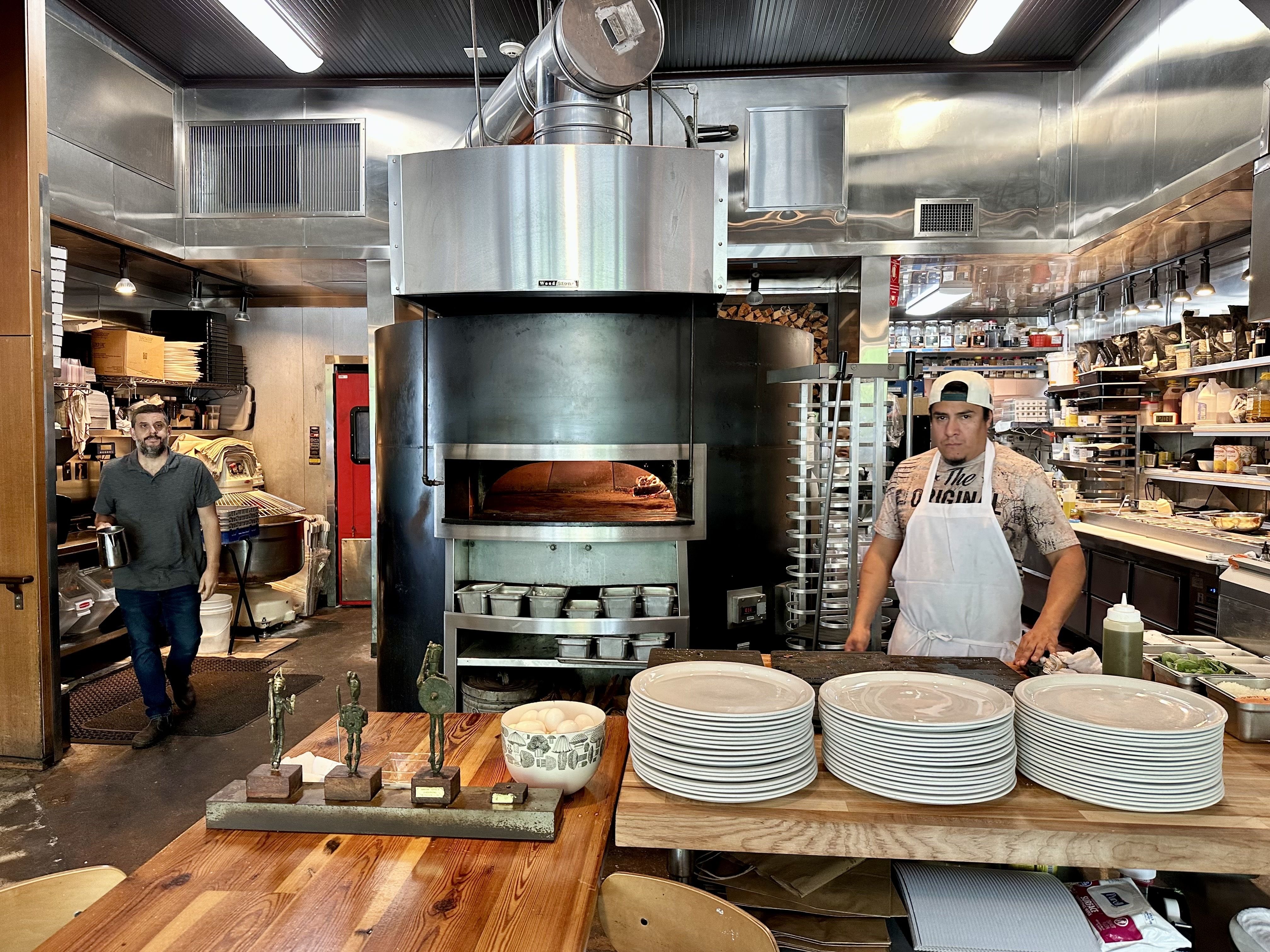 Industrial kitchen with a large round wood-fired oven and metal vent hood in the center. Two workers stand at the counter; stacks of plates and bowls on a wooden table and shelves of jars in the background.