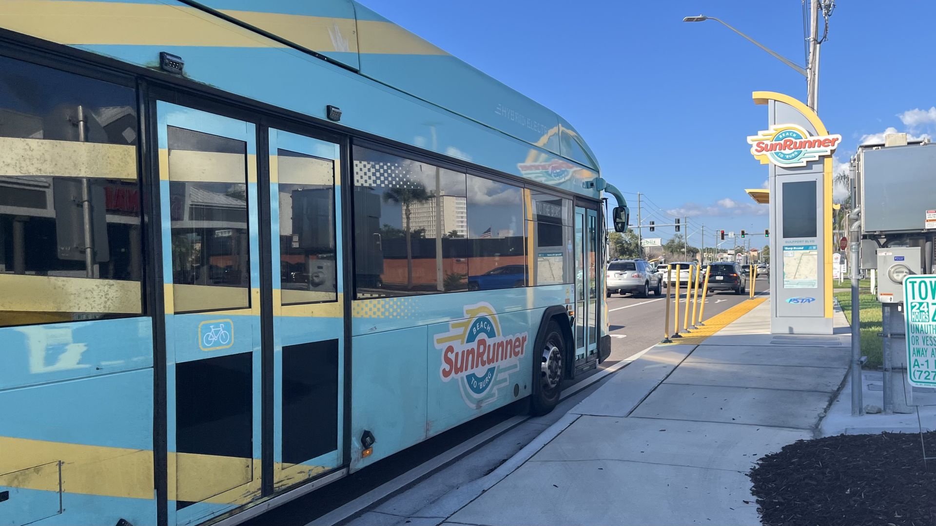 Turquoise and yellow SunRunner bus at a seaside stop with a prominent SunRunner sign, clear blue sky, cars in traffic, and a bike symbol on the bus door.