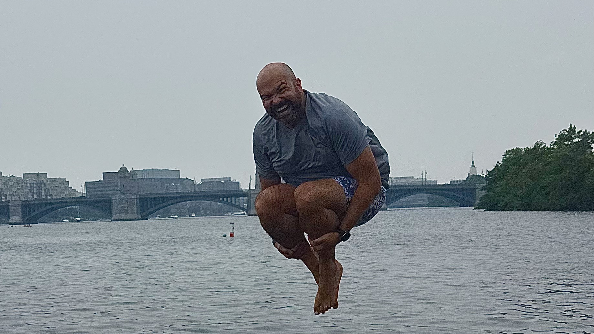 A bald man wearing a gray t-shirt and blue patterned shorts jumps mid-air above a river, with a city skyline, bridges, and trees in the background.