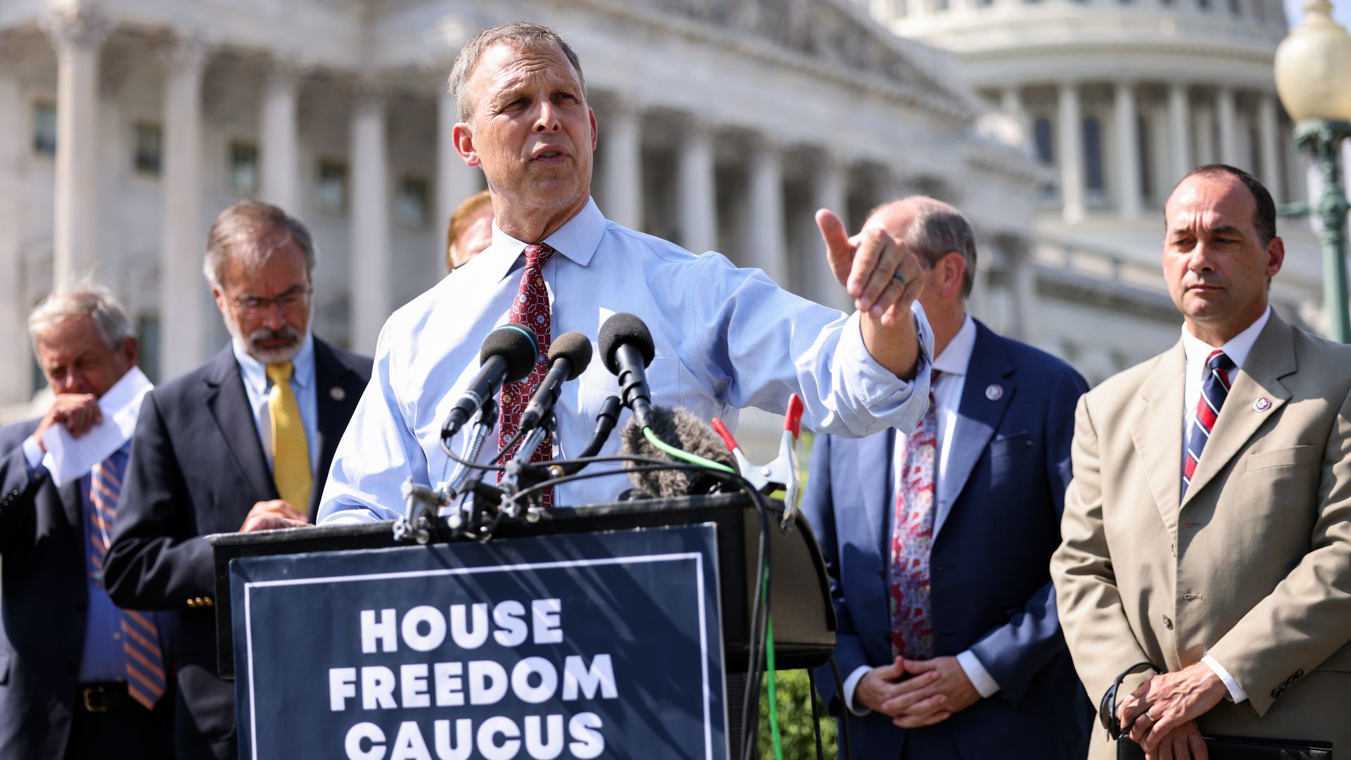 Man gestures while speaking at a podium that reads House Freedom Caucus in front of the U.S. Capitol building. 
