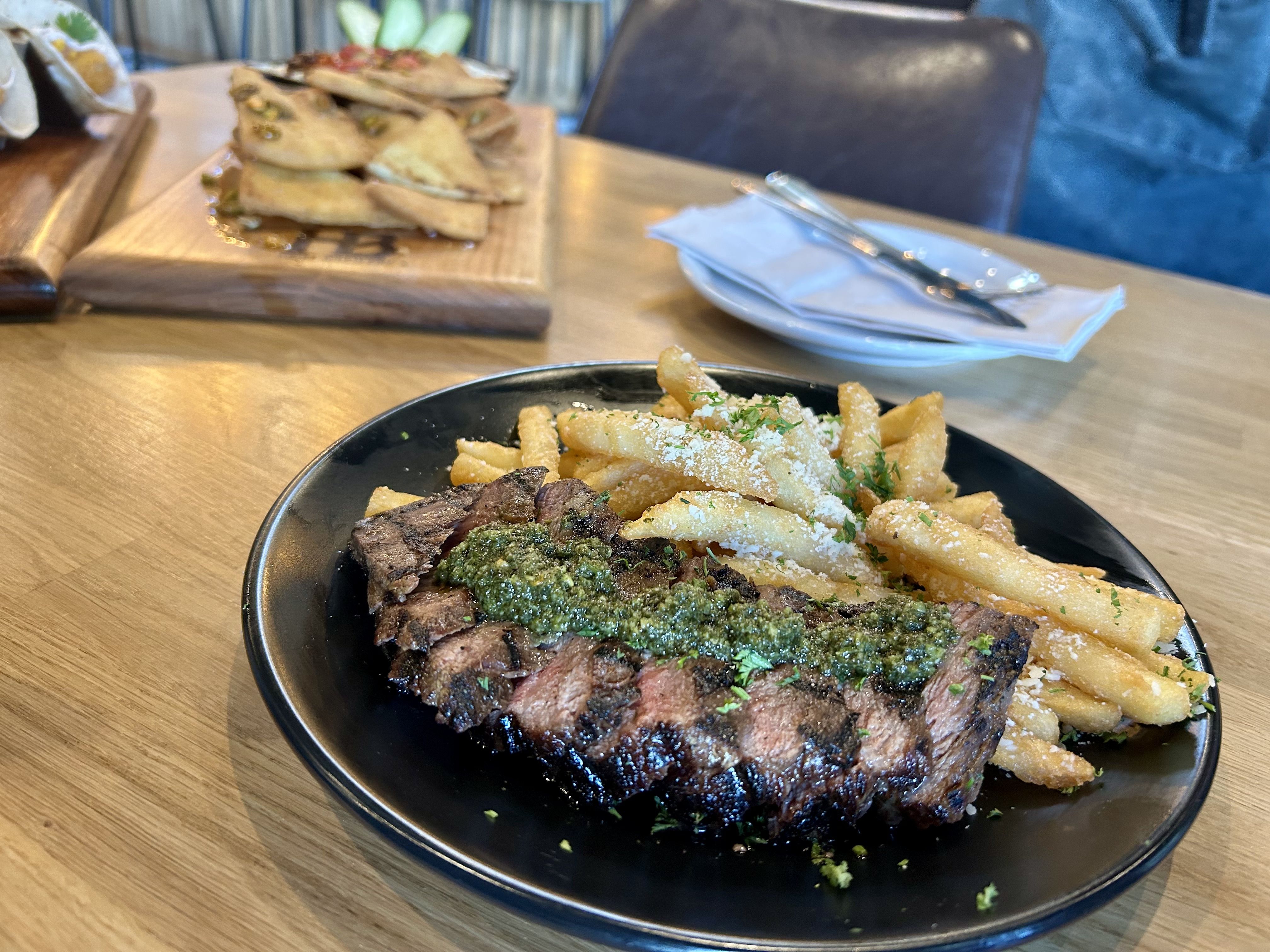 heist brewery. Black plate with sliced grilled steak topped by green herb sauce and fries, sprinkled with parmesan. Blurred background shows a wooden board with stacked pita sandwiches and a napkin with cutlery.
