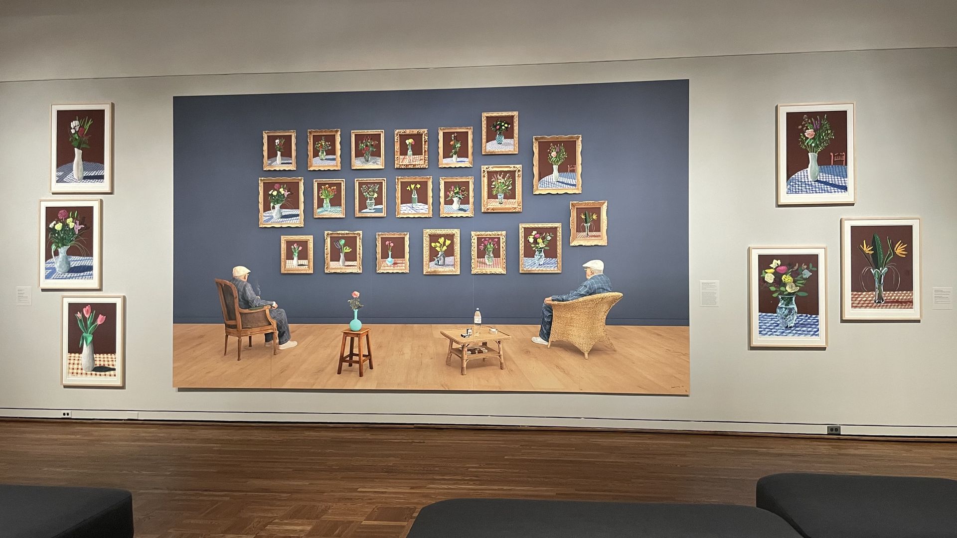 Art gallery wall featuring multiple paintings of flower vases on checkered tablecloths. Central large painting shows two elderly men seated facing a blue wall with various framed flower paintings.