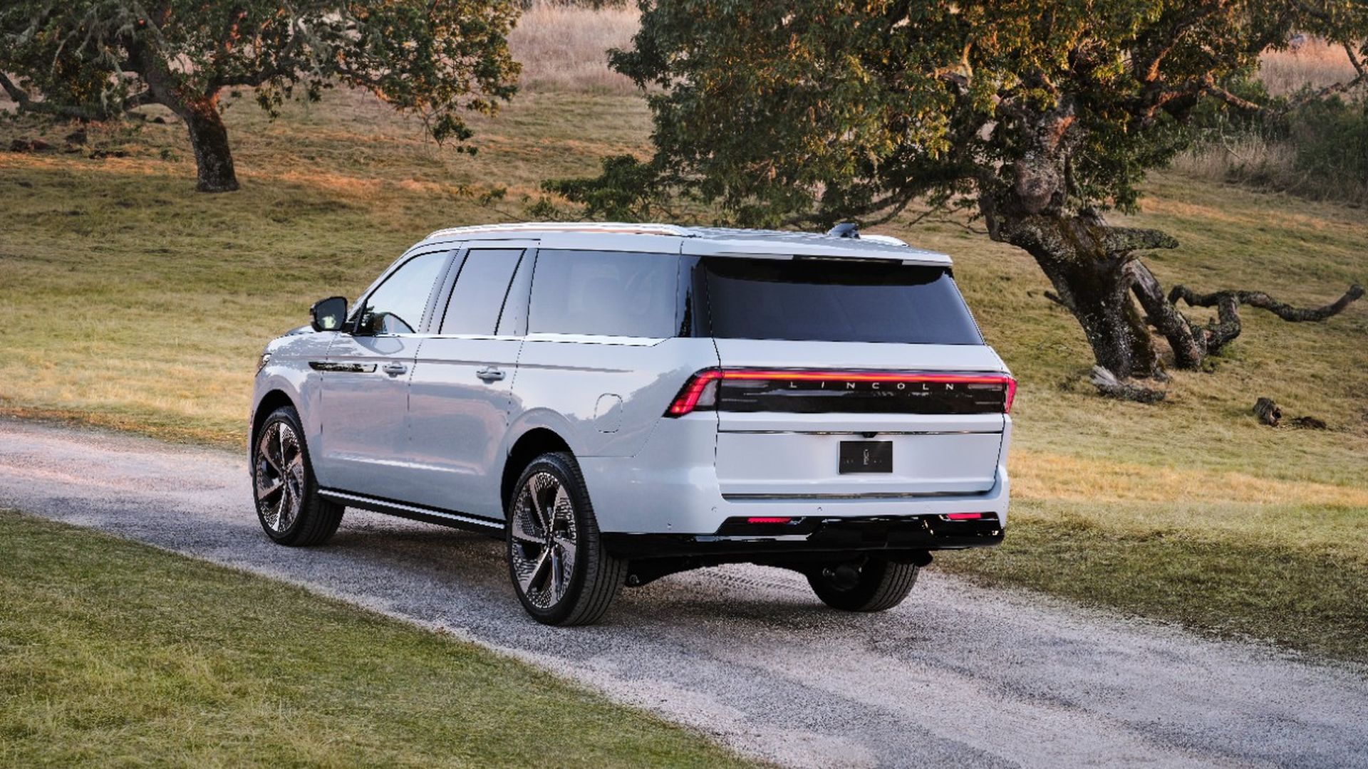 White Lincoln Navigator SUV on a gravel country road, rear three-quarter view, with grassy fields and large trees in the background.
