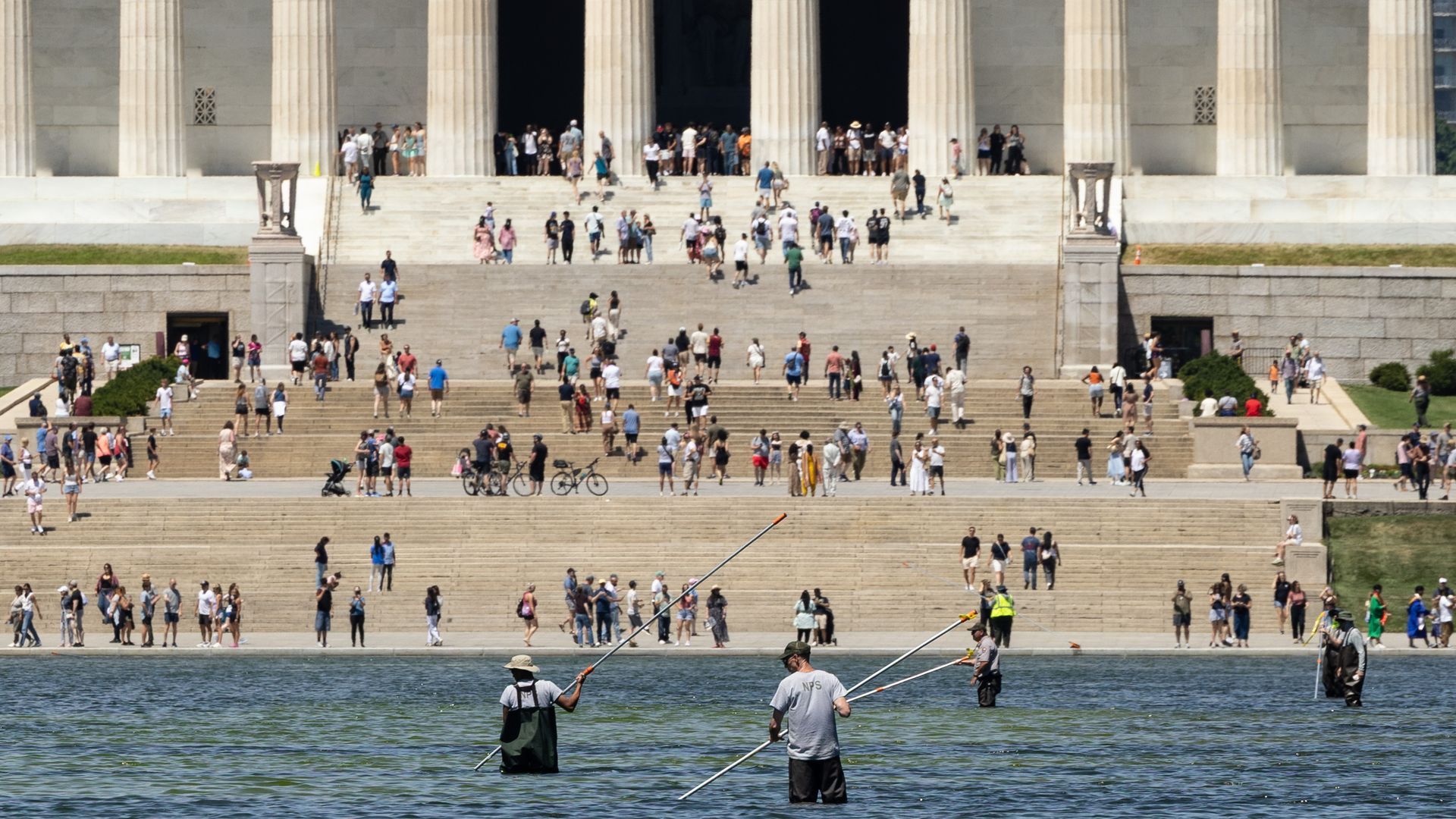 Wide view of a grand marble neoclassical building with tall columns and broad steps crowded with visitors; in the foreground, two fishermen in waders stand in water, casting their lines.