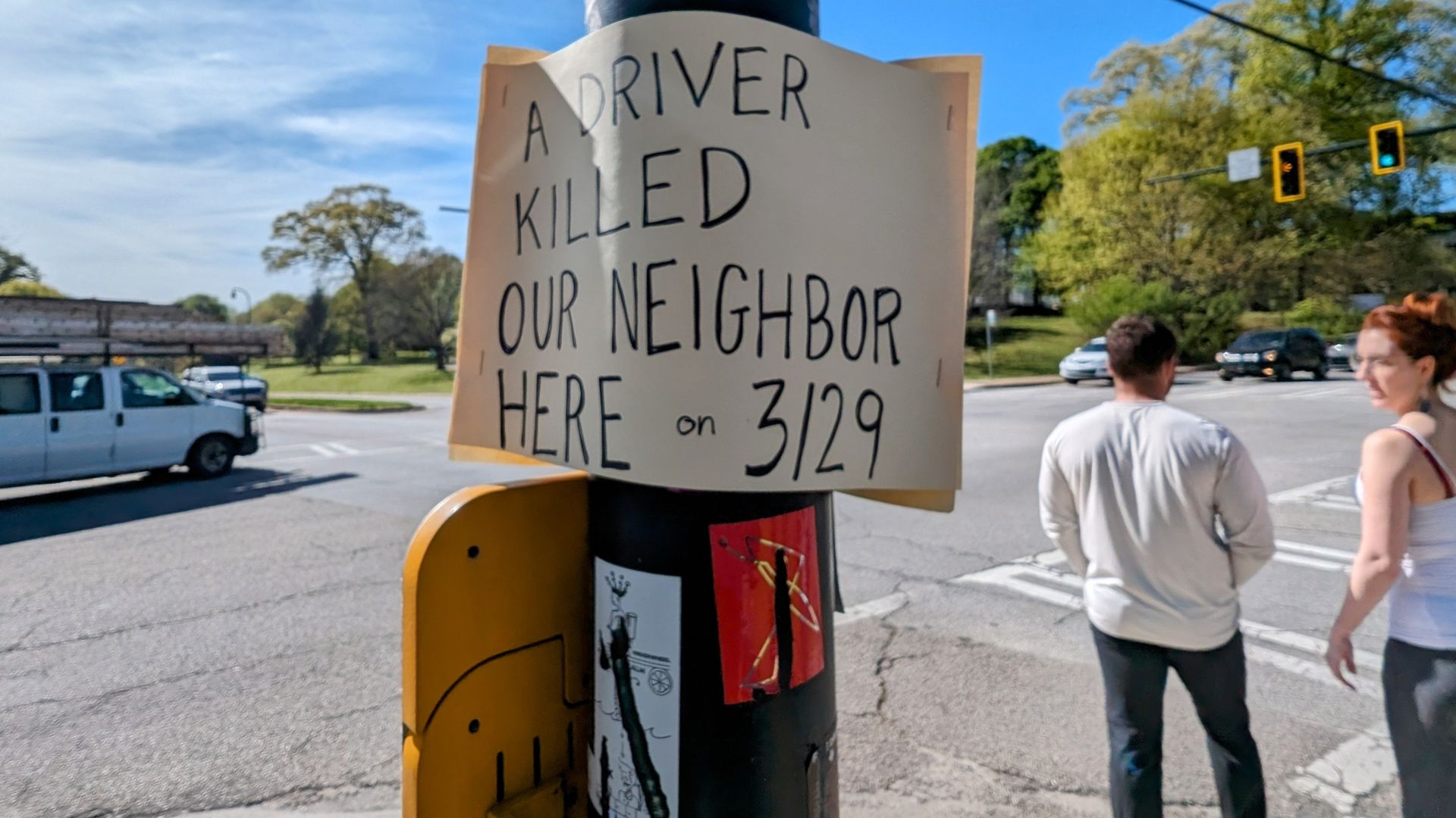 A handwritten sign on a black pole at a street corner reads: "A driver killed our neighbor here on 3/29." A white van and two pedestrians are visible near traffic lights.