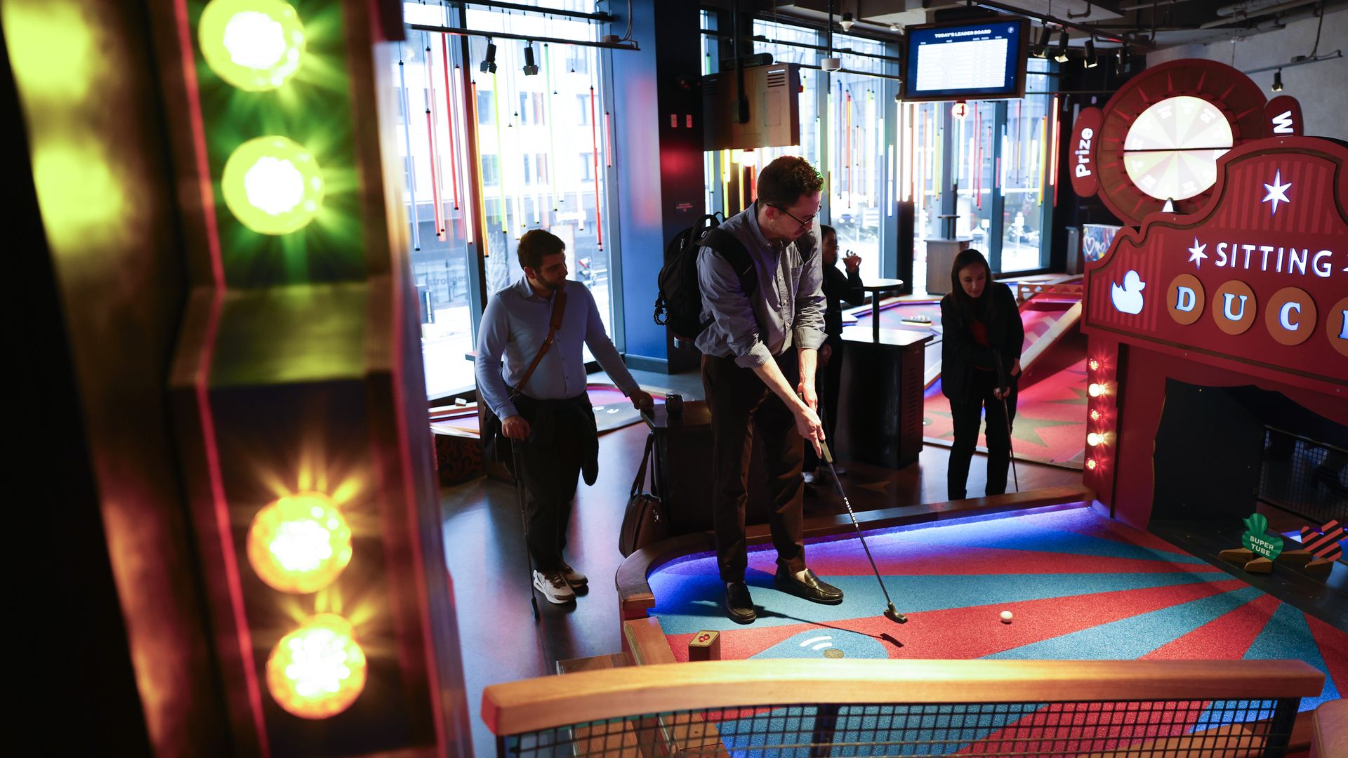 Indoor arcade mini-golf scene with neon colors: a man lines up a putt as two others watch; bright red "SITTING DUCK" sign and a large wheel on the right.