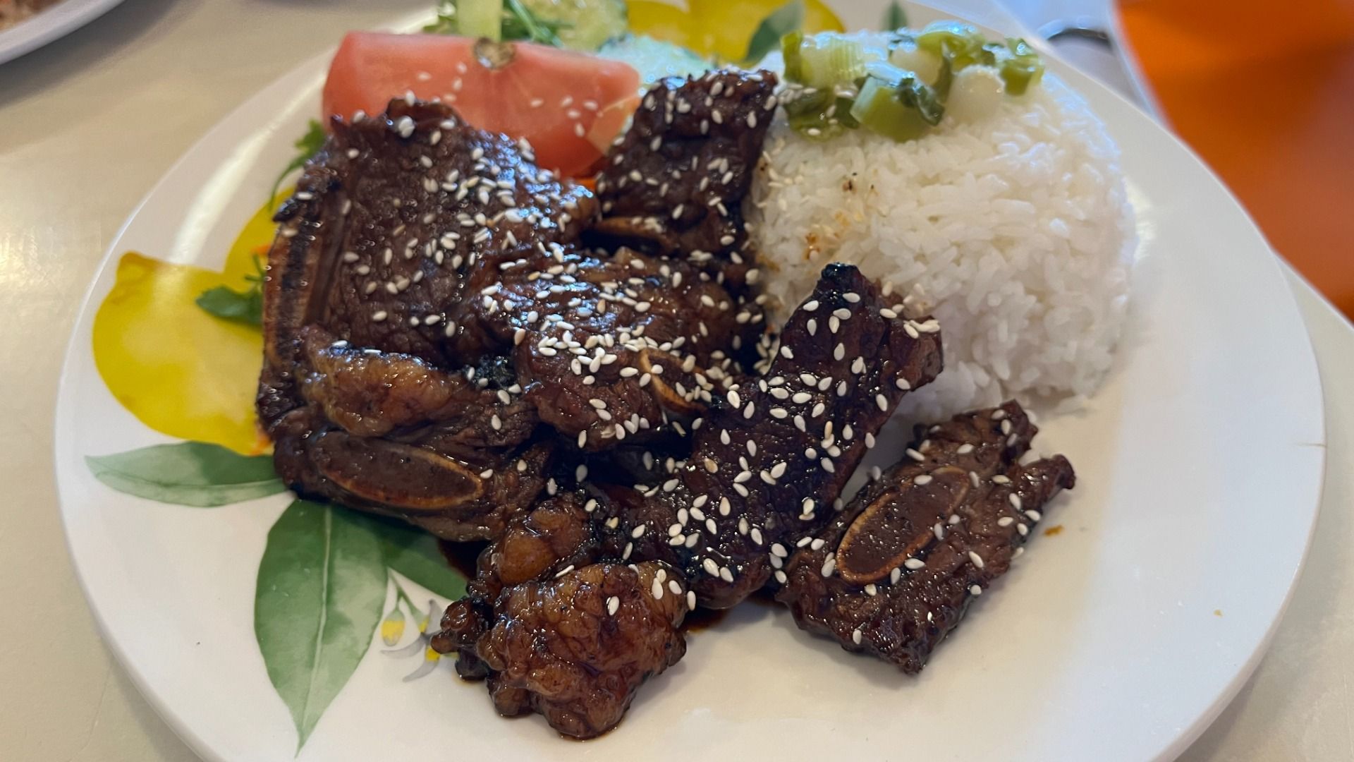 Glazed beef ribs coated with sesame seeds atop a plate, beside a mound of white rice with chopped green onions, a tomato slice, and a yellow sauce smear on a decorative plate.