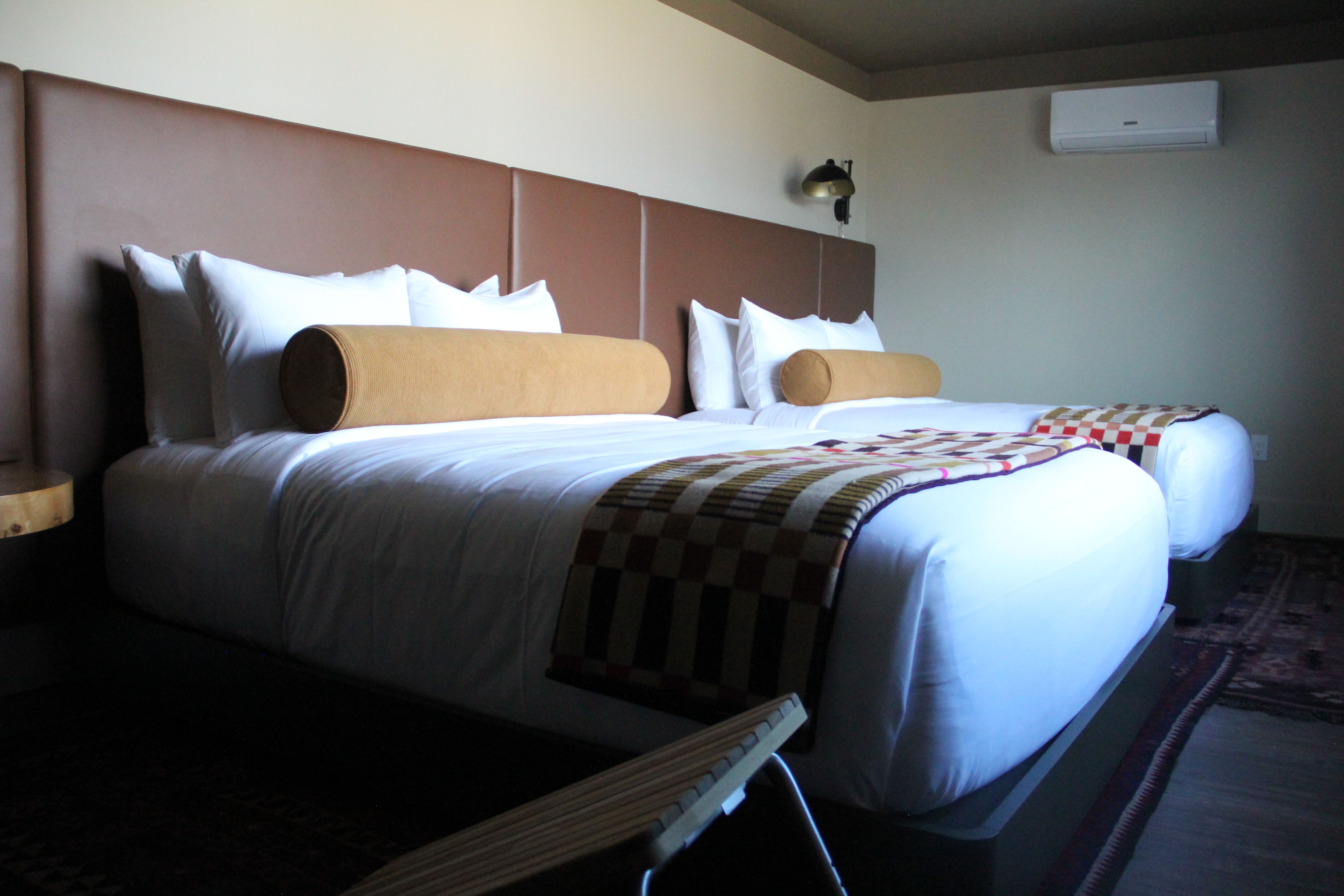 Two large white beds with tan padded headboards and cylindrical beige bolsters. A checkered throw drapes the foot; wall lamps, an air conditioner, and a patterned rug complete the room.