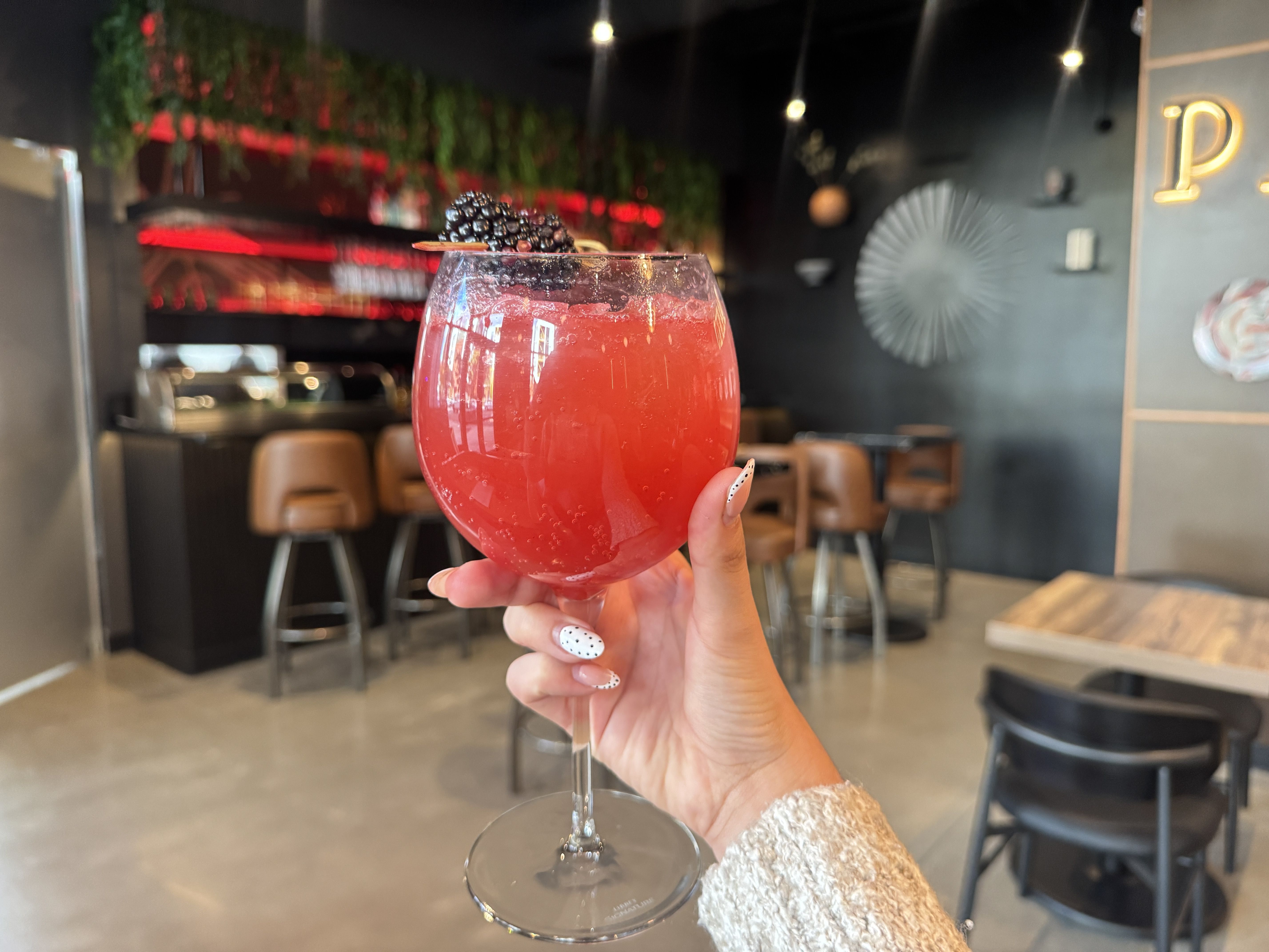 Hand holding a large glass with a pink-red drink garnished with blackberries on a skewer, in a modern bar with brown stools and dark walls with red lighting accents.