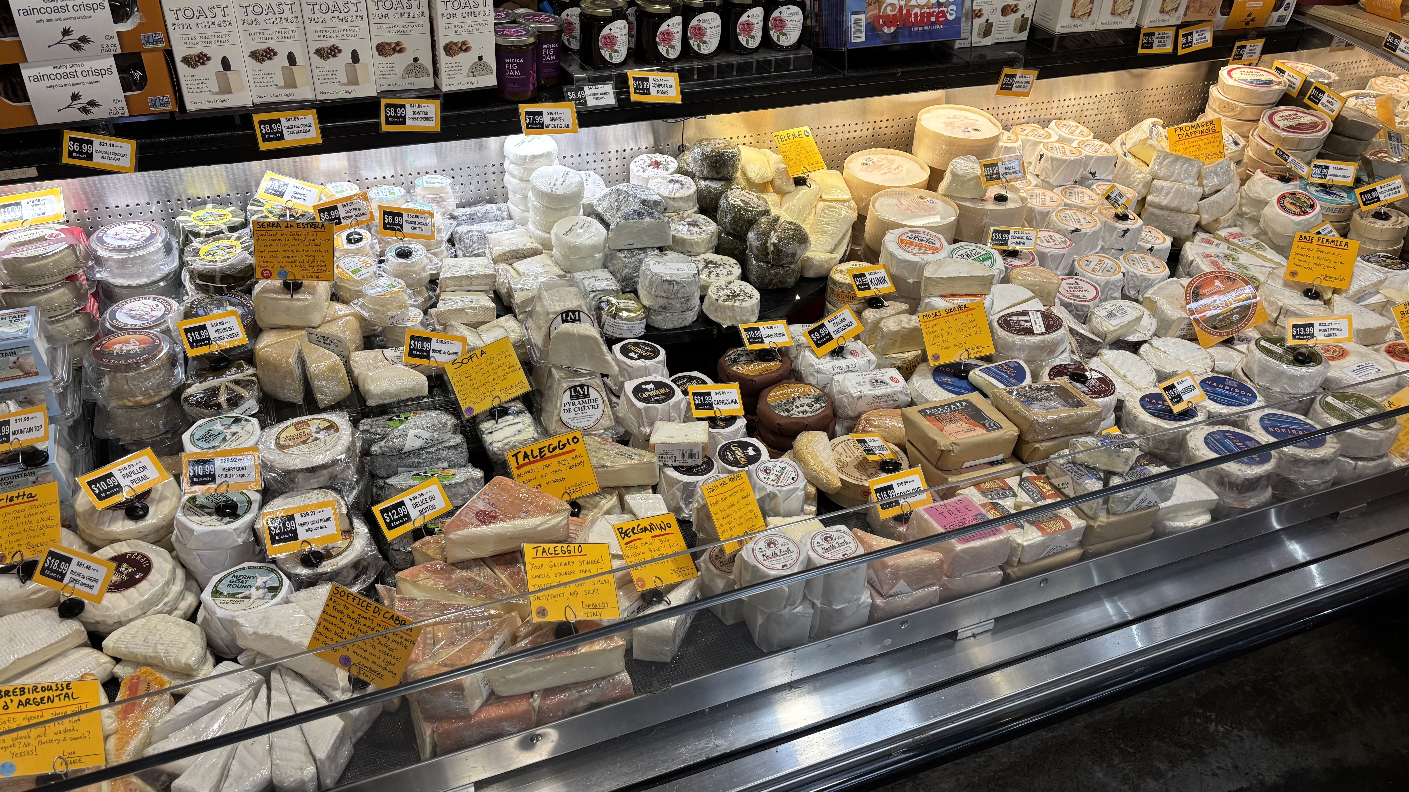 Display case with various cheeses in white, beige, and yellow tones, some wrapped, others round or wedge-shaped, with price tags and descriptions on yellow and white cards, plus crackers on top shelf.