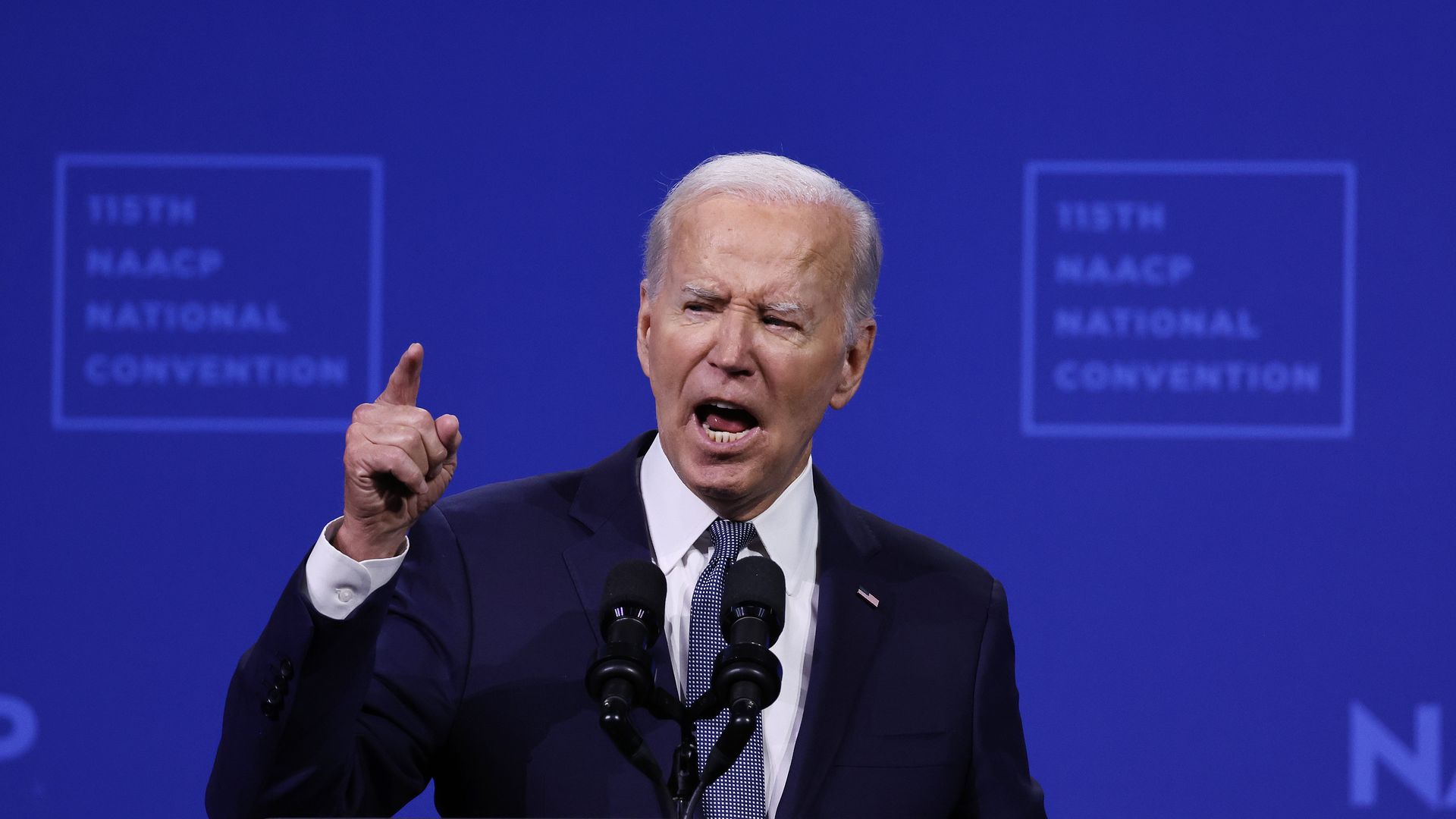 President Biden, holding up his pointer finger in behind a microphone and in front of a blue NAACP backdrop.