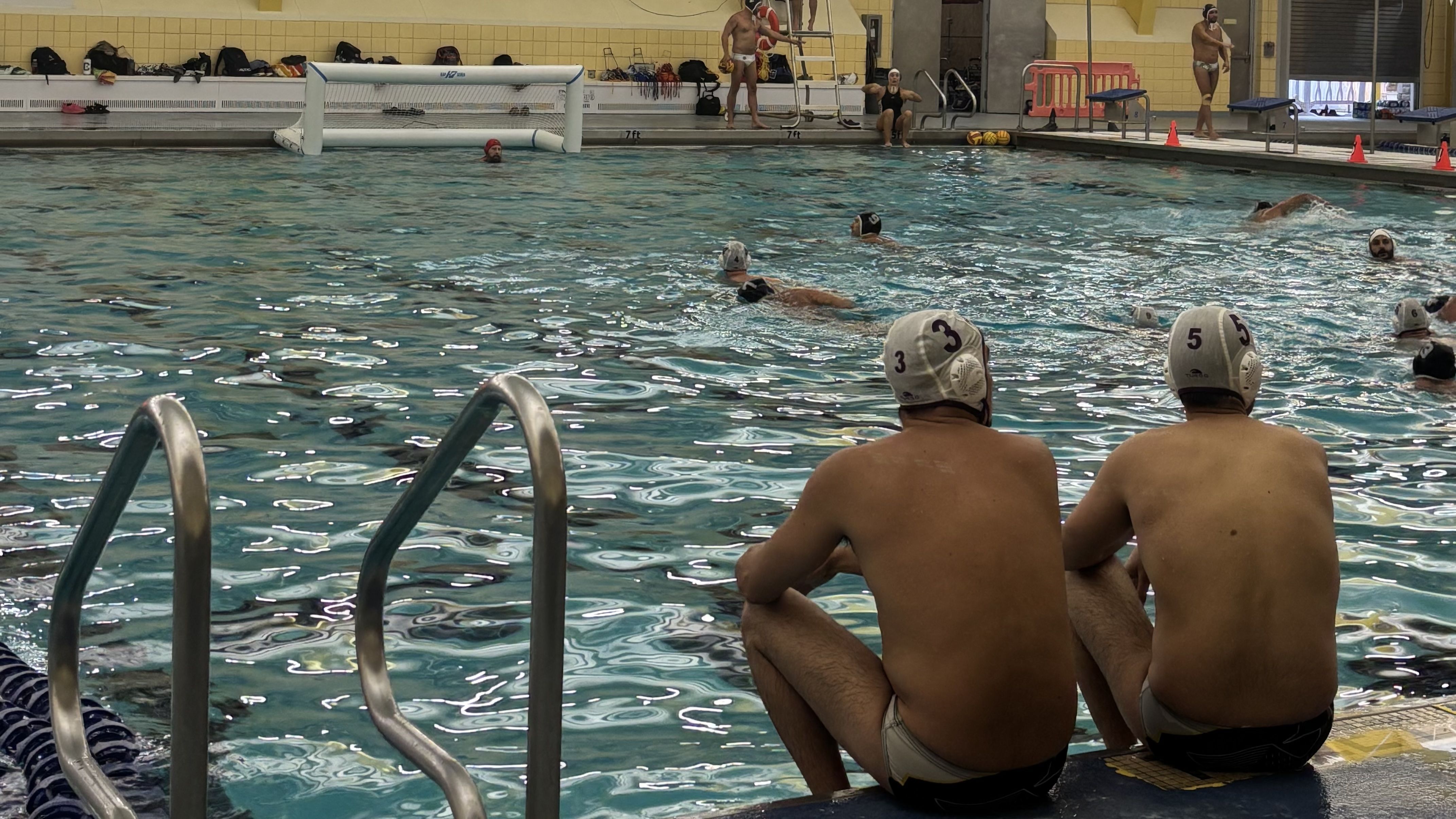 Two men sit at the edge of a pool while watching a water polo game.