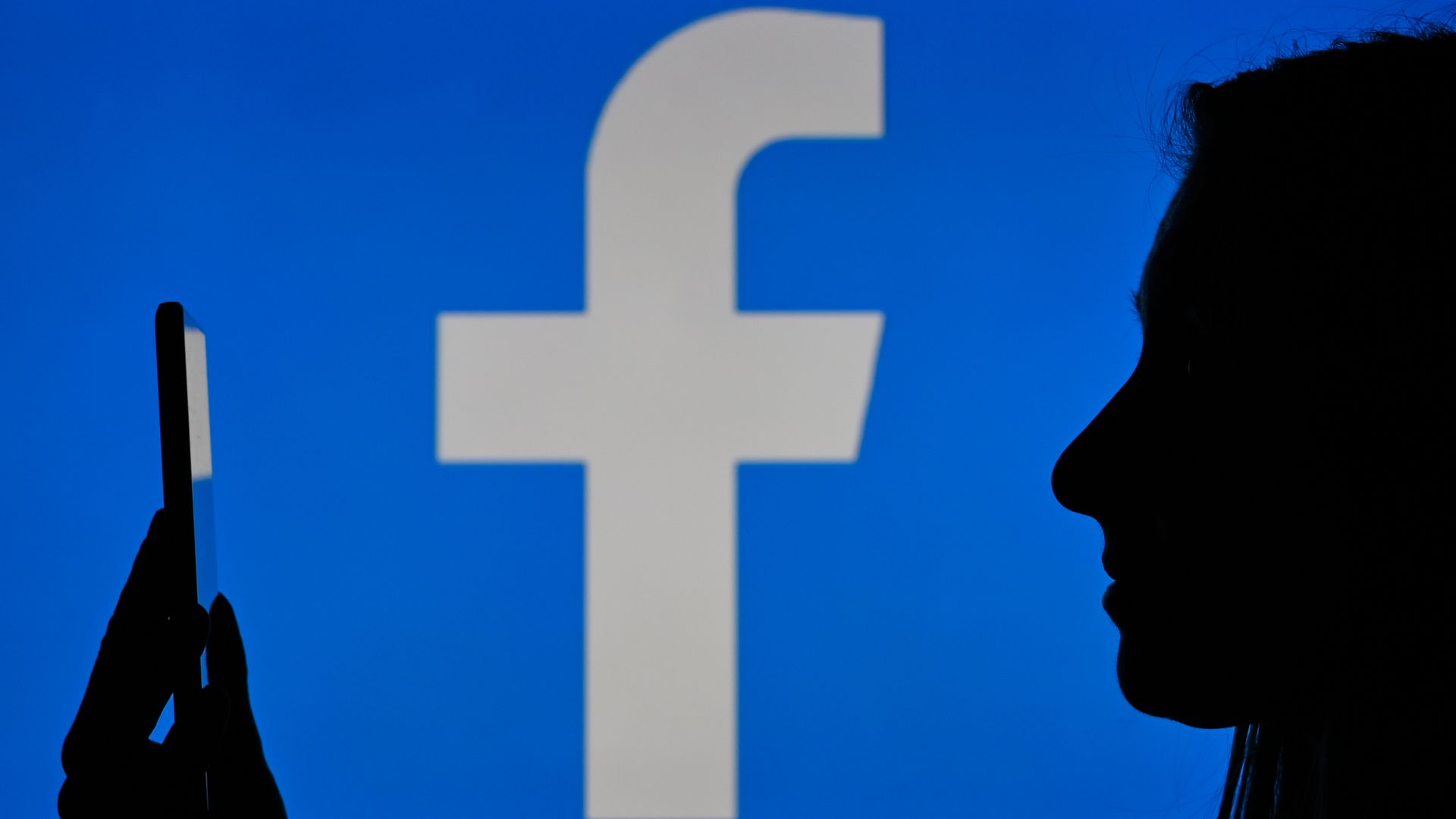 An image of a woman holding a cell phone in front of a Facebook logo displayed on a computer screen. 