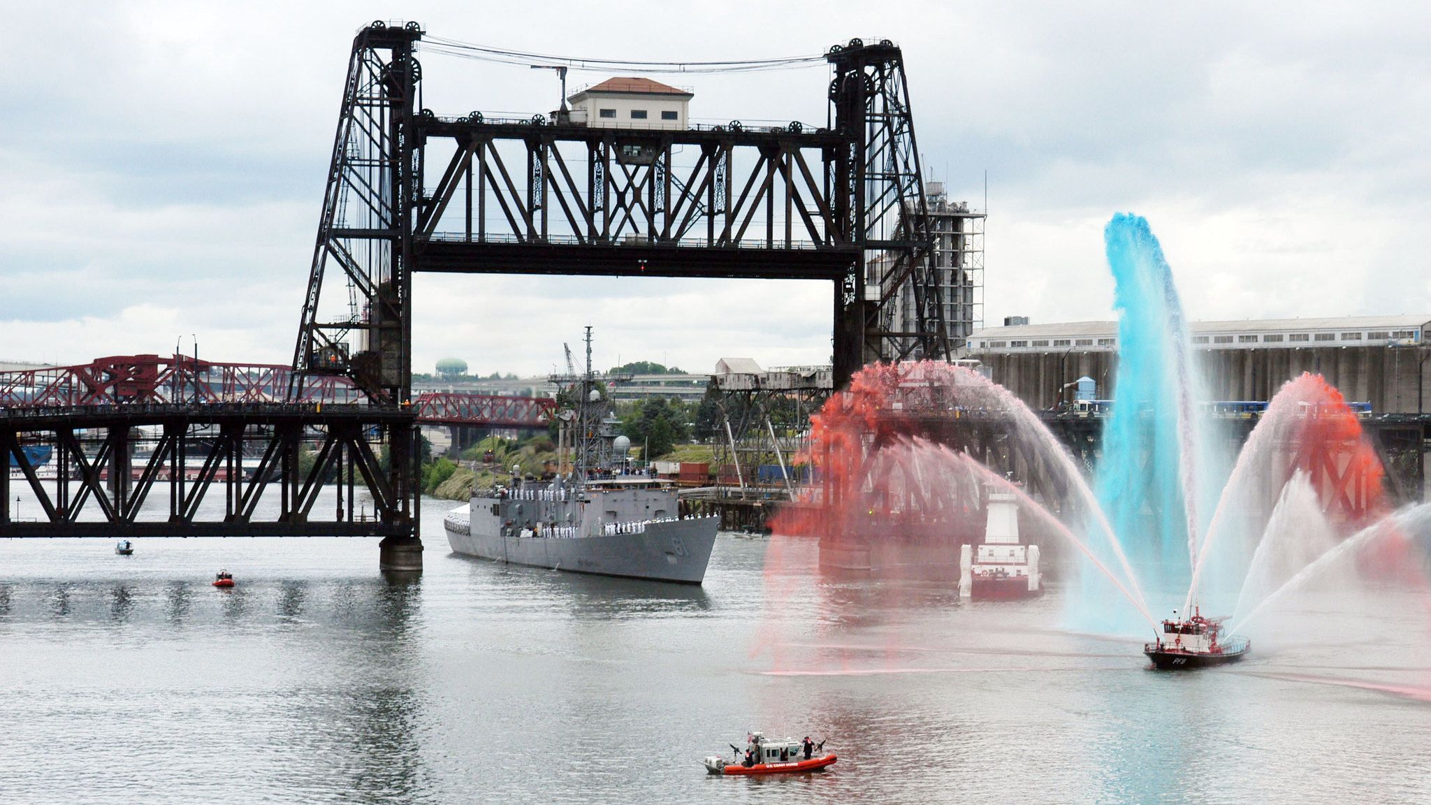 Large dark steel vertical-lift bridge spans a river; a gray warship passes underneath. In the foreground, red and blue fountain jets arc across the water, with small boats nearby.