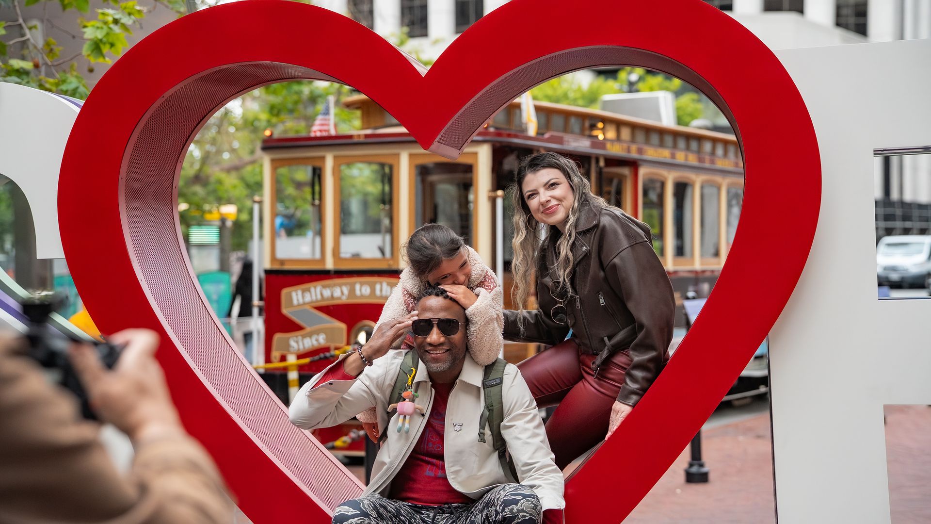 A diverse family posing happily inside a large red heart-shaped frame with a vintage cable car and urban buildings in the background during daytime.