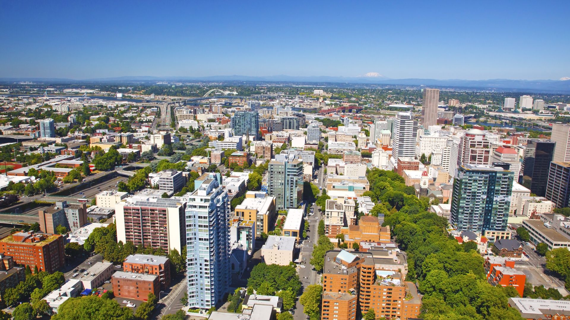 Aerial view of a city with numerous buildings, green trees lining streets, a clear blue sky, and mountains in the far background under bright daylight.