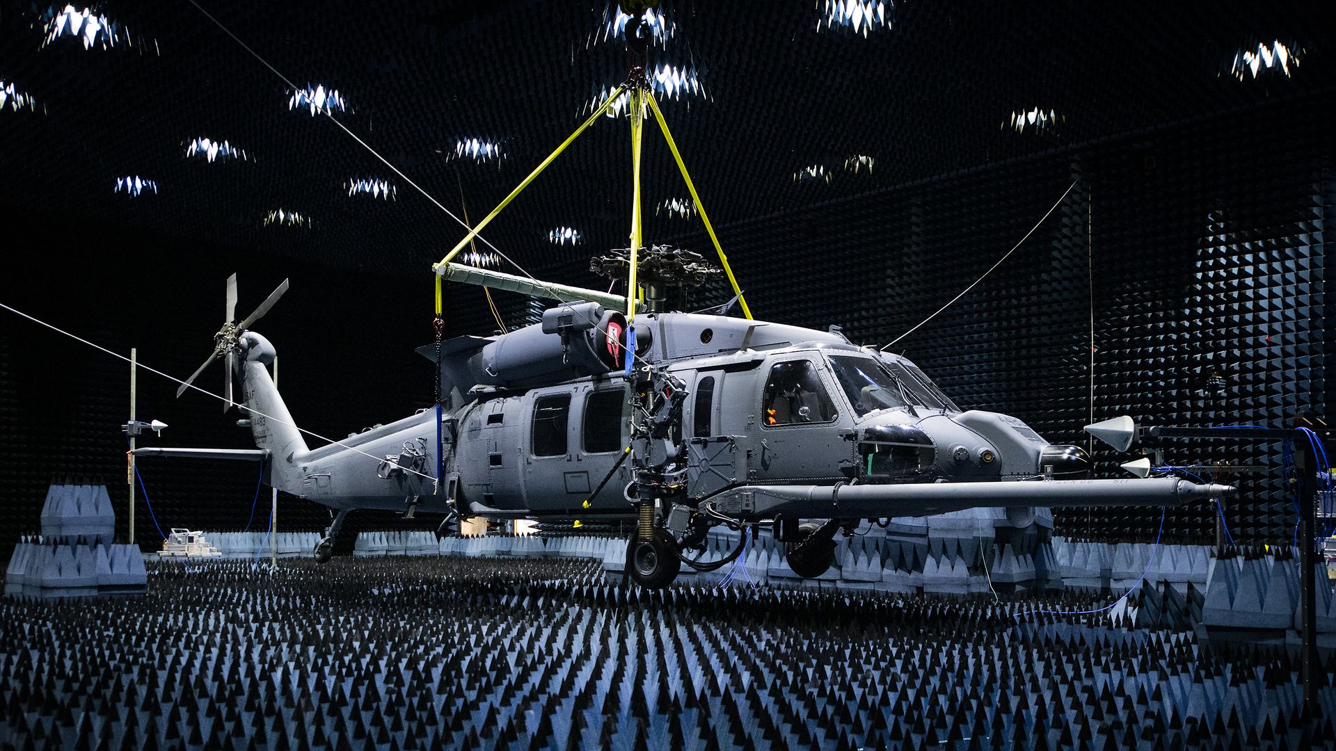 A large, grey helicopter is suspended in an anechoic chamber. Yellow straps hold it up. Protrusions from the wall are dark in color, mostly blue and black.