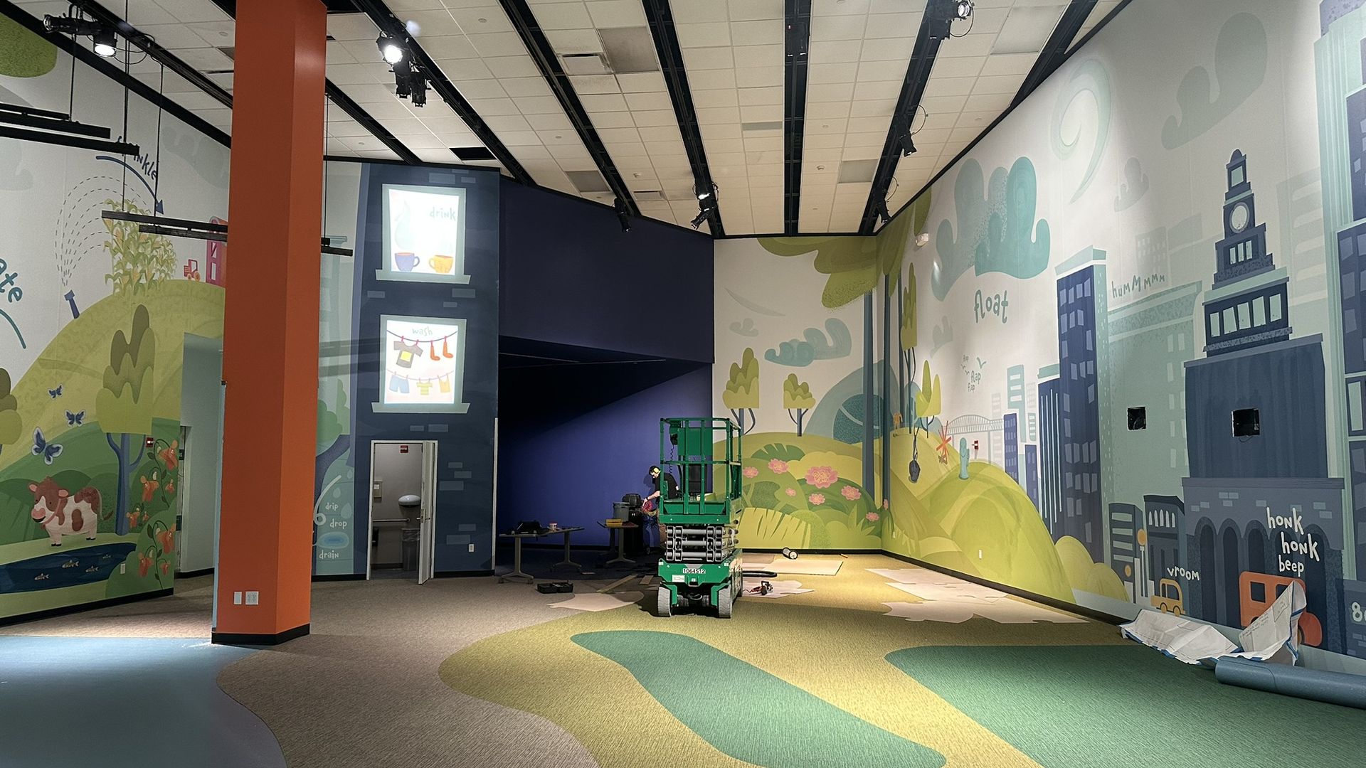 Indoor space with colorful murals of nature, animals, and a cityscape on walls, a tall orange pillar, green scissor lift, and multicolored carpeted floor in shades of green, brown, and blue.
