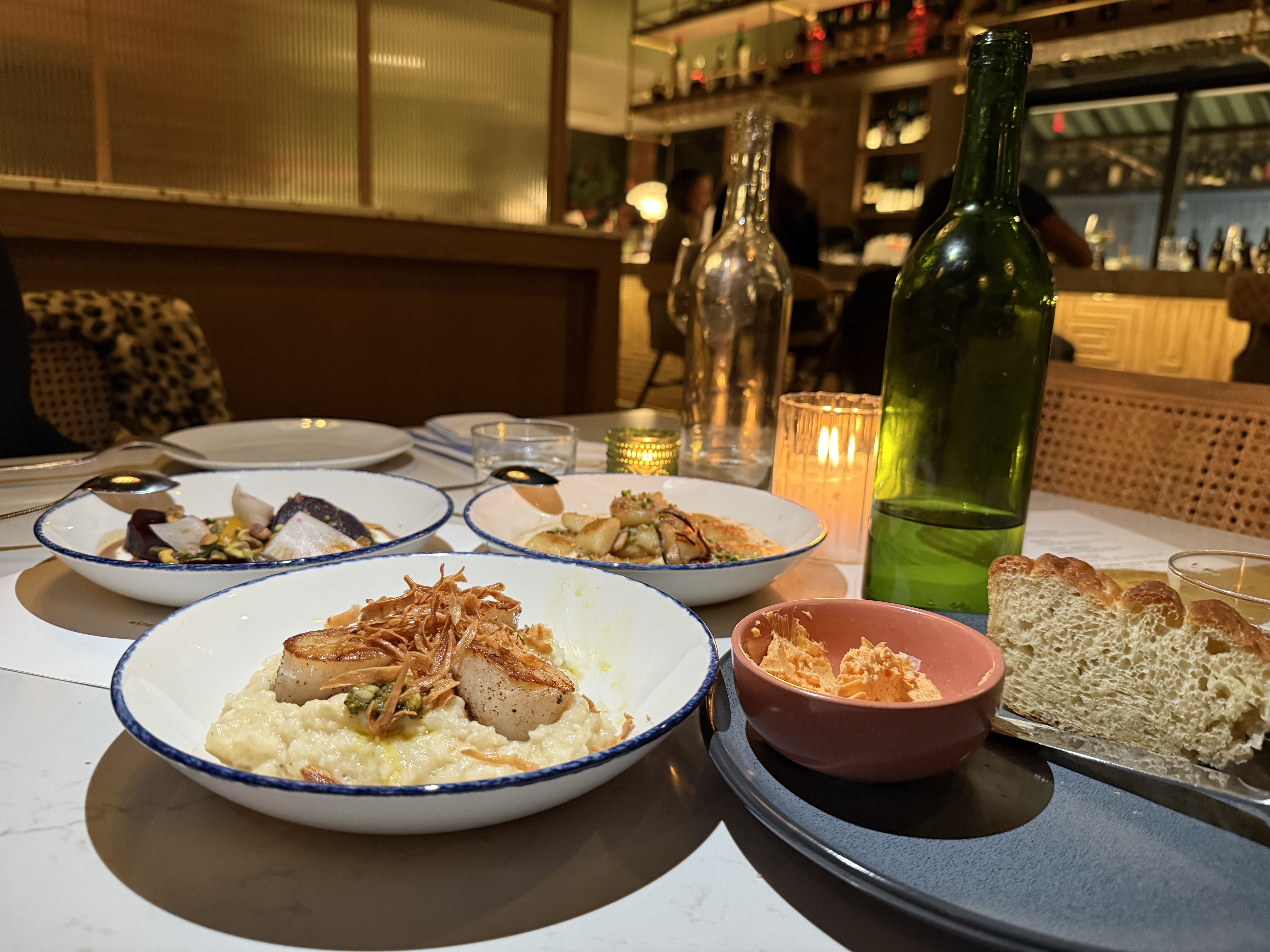 Table setting in a cozy restaurant with three dishes: seared scallops on risotto, mixed roasted vegetables, and another plate with shrimp. Also bread with spread, a green glass bottle, and candlelight.