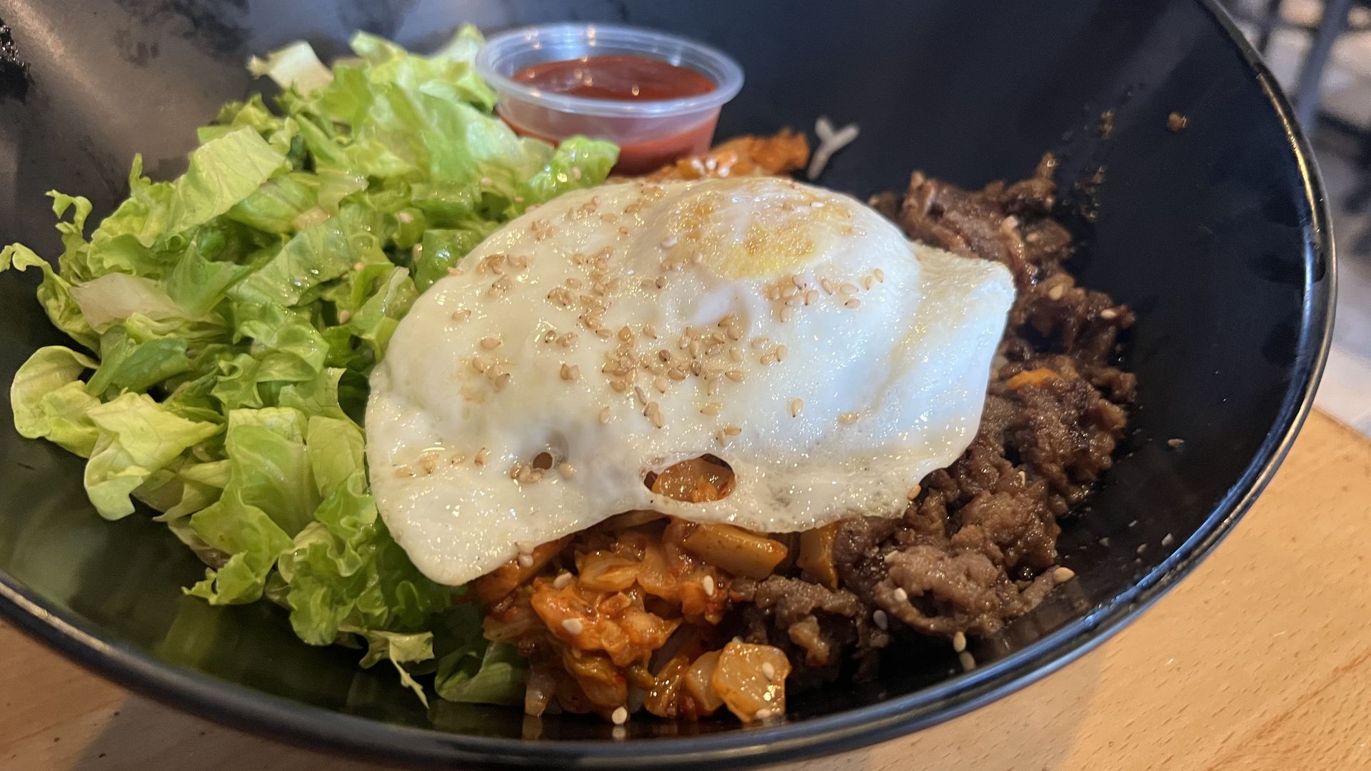 Black bowl with green lettuce, fried egg sprinkled with sesame seeds, seasoned cooked beef, and reddish kimchi, alongside a small container of red sauce on a light wooden surface.