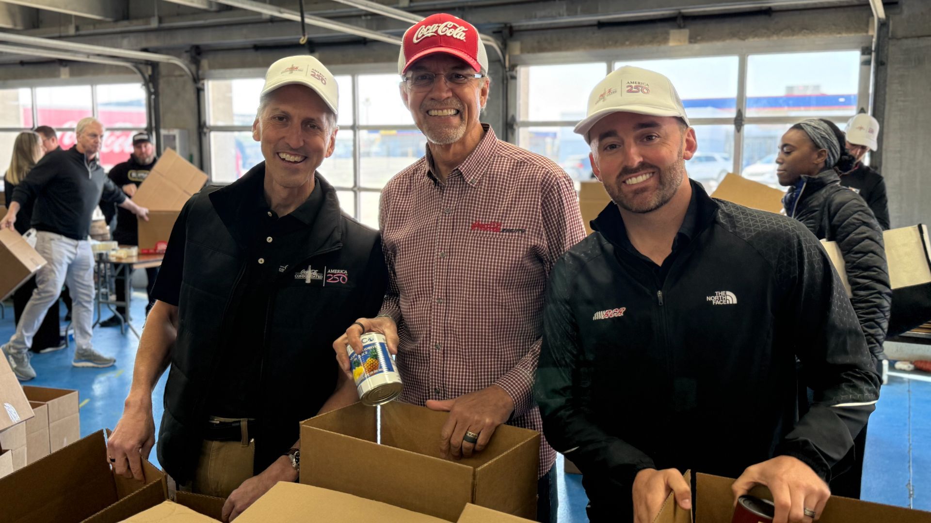 Three men smile at a packing station during a donation drive; center man wears a red Coca-Cola cap and checkered shirt, flanked by others in white caps and dark jackets, packing boxes with cans.