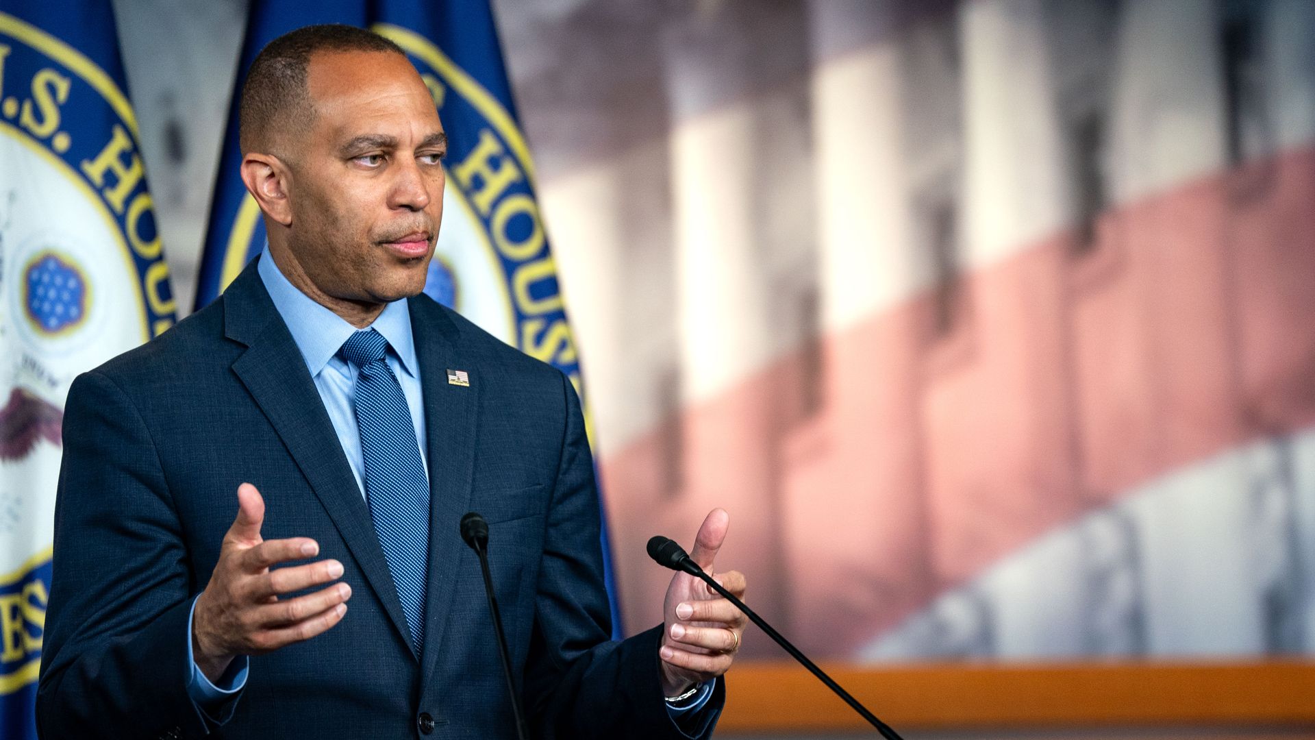 House Minority Leader Hakeem Jeffries, wearing a blue suit with his arms raised at his sides, standing behind microphones and in front of two House of Representatives flags, as well as a mural with an opaque American flag transposed over the Capitol.