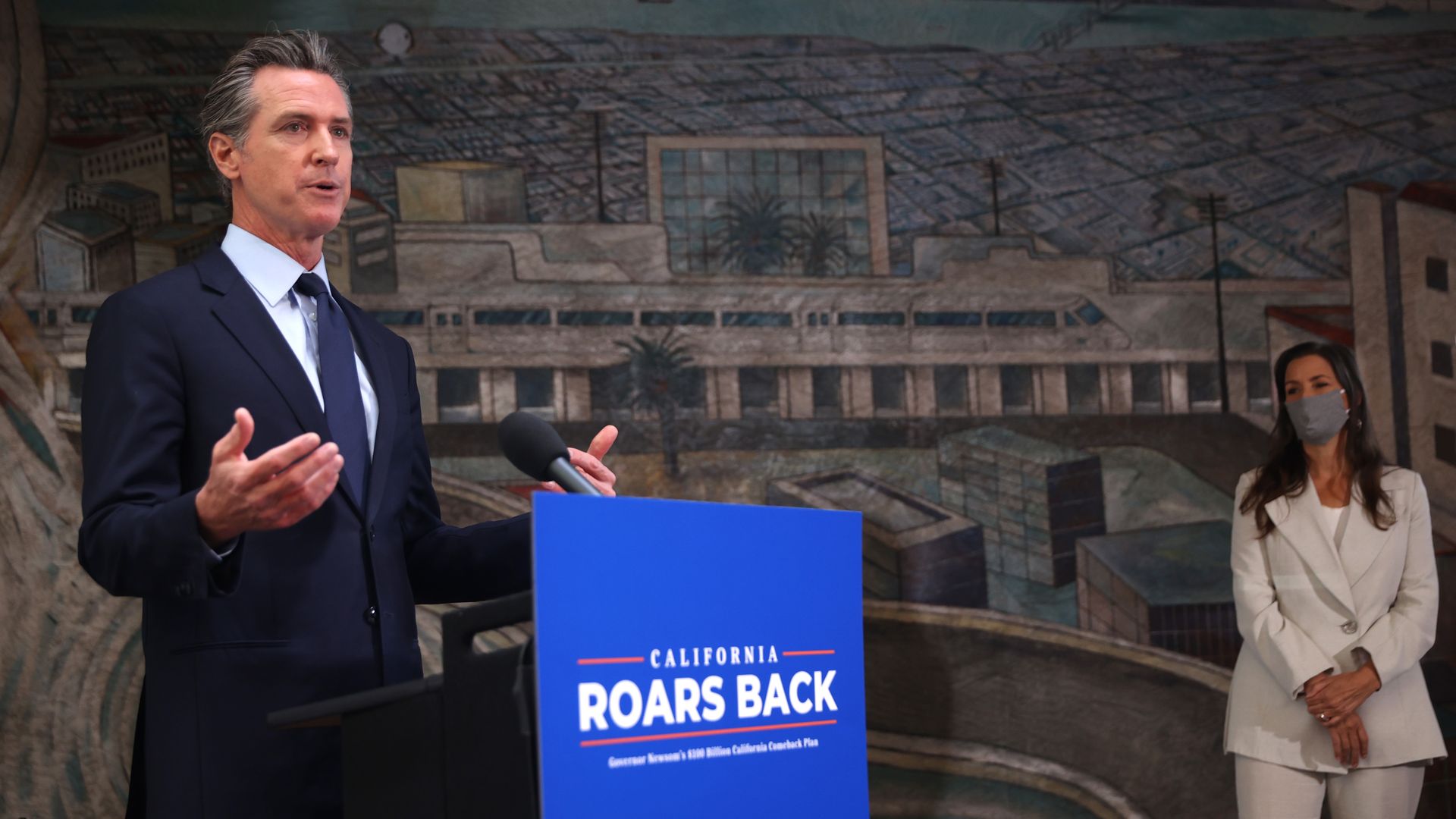 Photo of Gavin Newsom speaking while standing next to a podium that has the sign "California roars back"