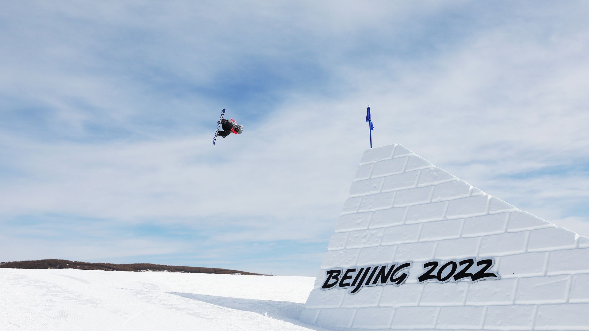 A snowboarder performs an aerial trick in a snowy landscape under a cloudy sky, next to a snow ramp with "Beijing 2022" written on it and a blue flag on top.