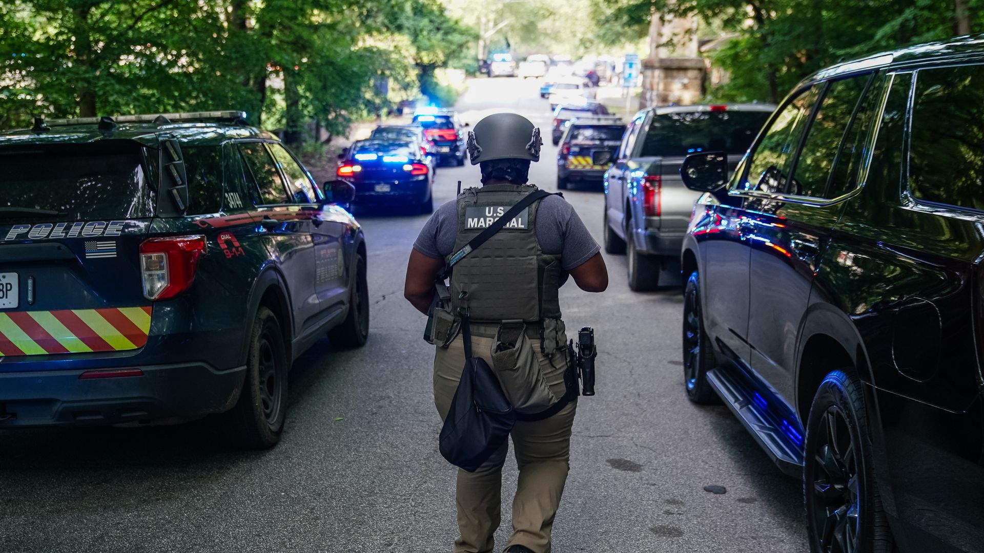 A U.S. Marshal wearing a bulletproof vest and helmet walks between parked police vehicles with flashing blue lights on a tree-lined street during a law enforcement operation.