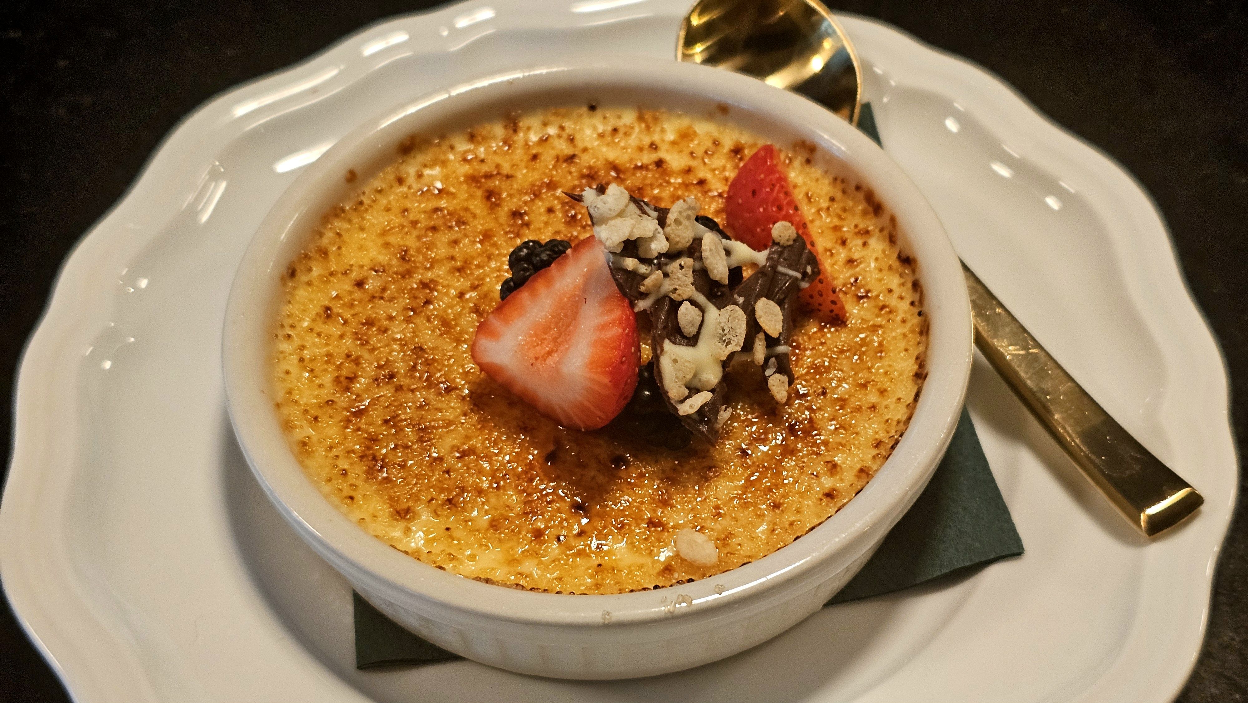 Crème brûlée with a caramelized top, garnished with strawberry halves, chocolate shavings, and crushed nuts, served in a white ramekin on a white plate with a gold spoon.