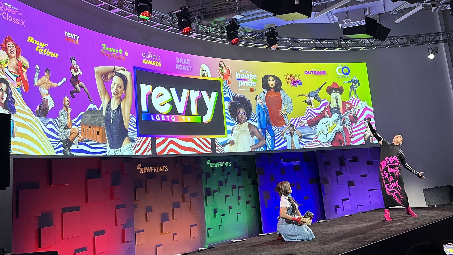 One person dressed as Dorothy from the Wizard of Oz and other in a black dress that says "Drag is not a crime" standing in front of a rainbow lit backdrop with Revry on a screen