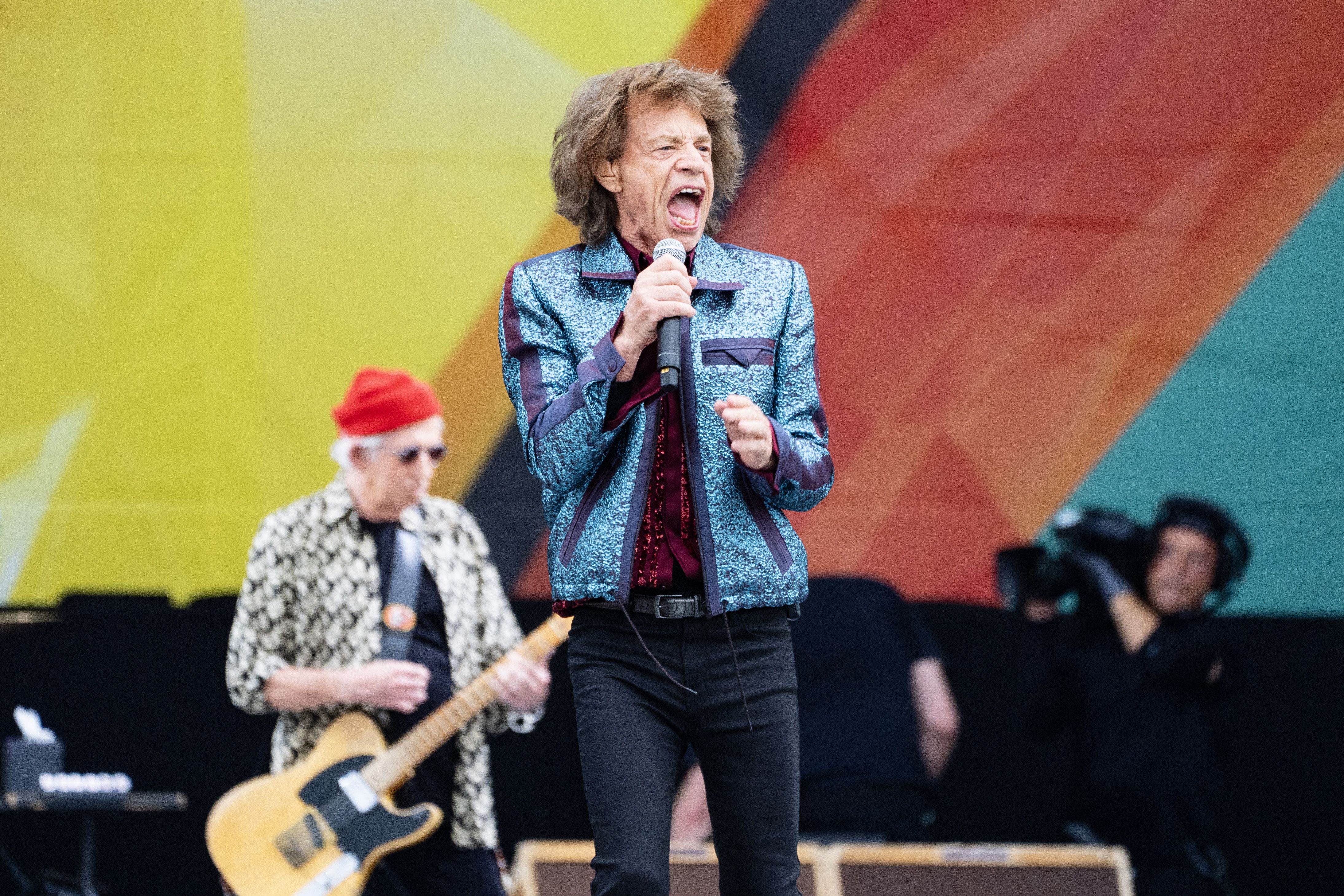 Mick Jagger sings into a microphone. His hair is lifted as though he is mid-jump.