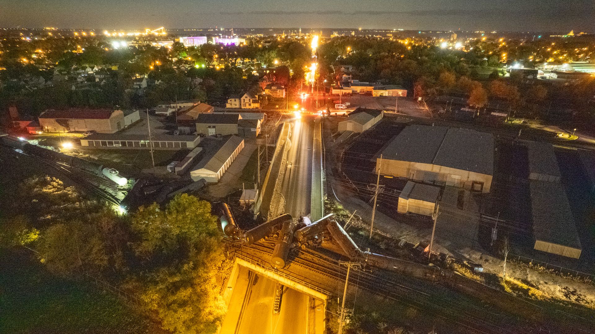 Derailed train cars on an underpass bridge, with evening lights from a lakefront town in the distance
